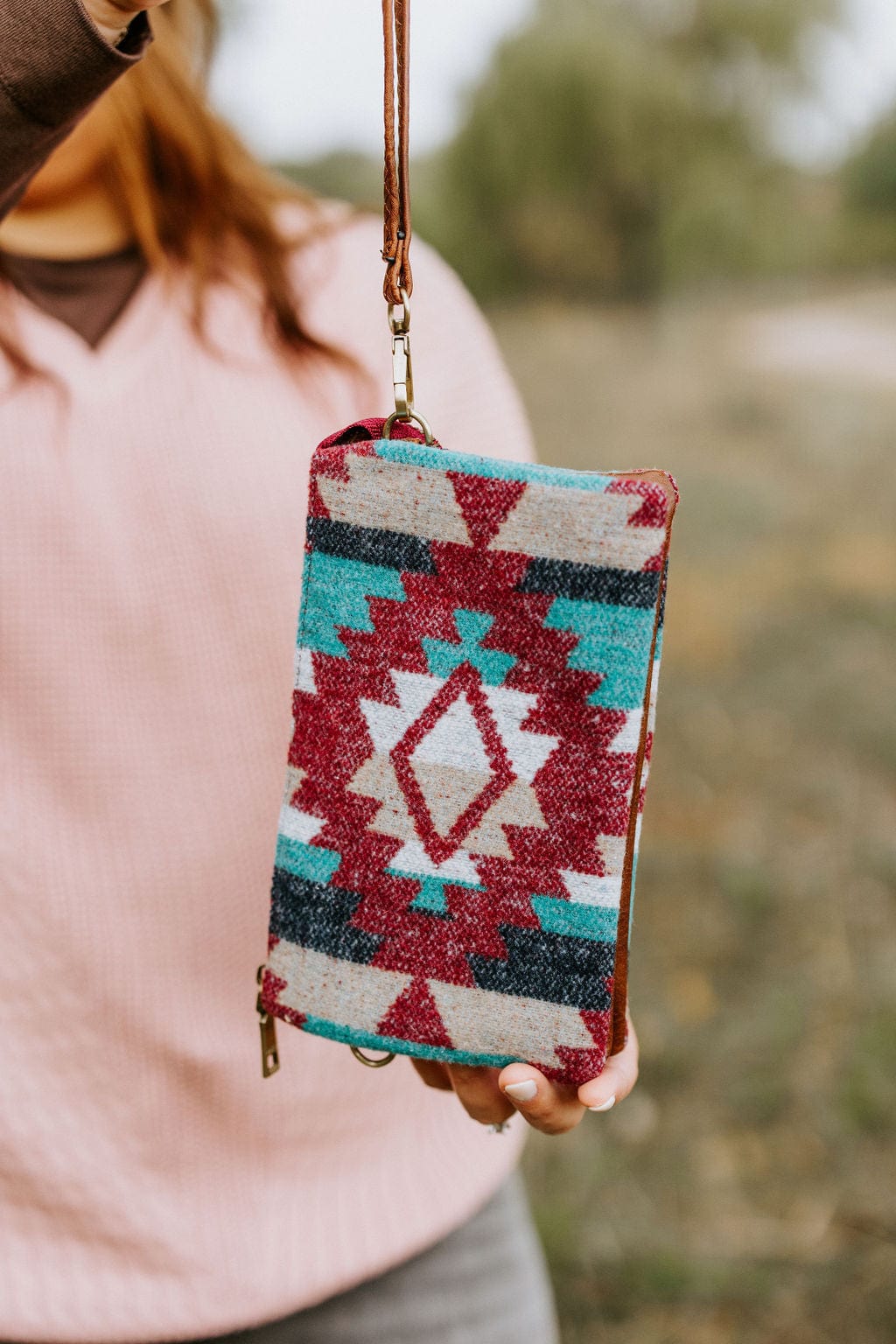 A person holding a vibrant AZTEC CLUTCH, adorned with geometric designs in red, teal, and beige tones. The clutch features an adjustable crossbody strap and faux leather detail, adding a touch of versatility and style.