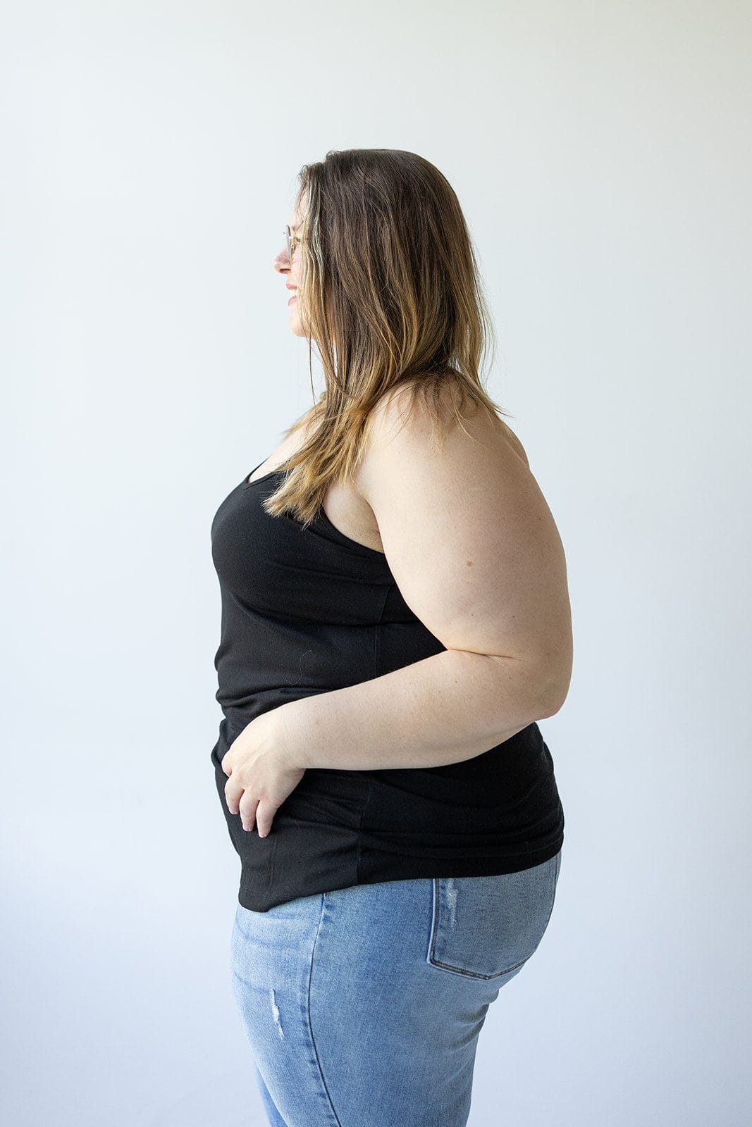 A person with long hair and glasses is wearing the BASIC RACERBACK CAMI IN BLACK by Love Marlow, paired with blue jeans, standing in profile against a plain white background.