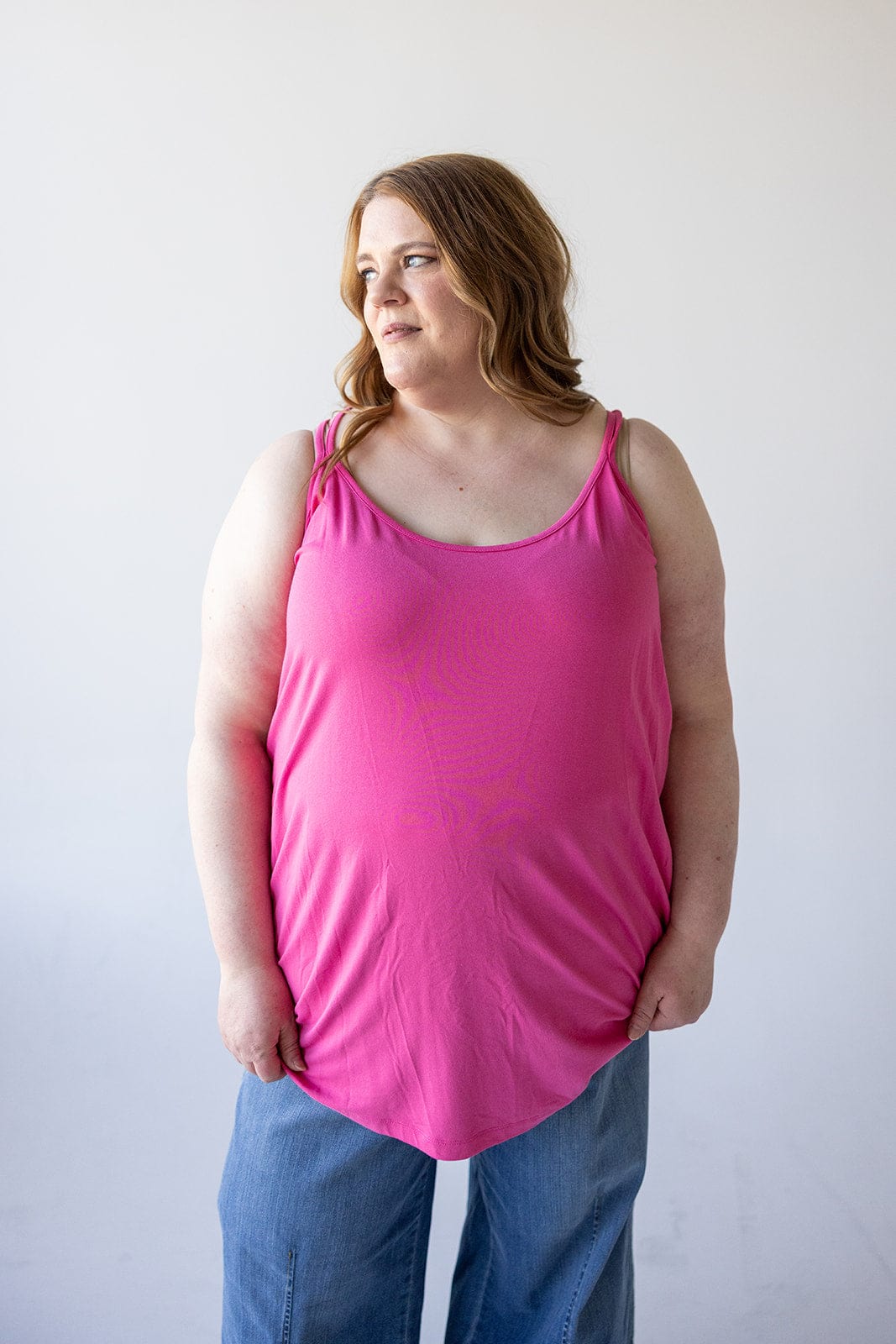 A person with long hair, wearing a Love Marlow BASIC RACERBACK CAMI IN FRESH FUCHSIA and blue jeans, stands against a plain white background looking to the side.