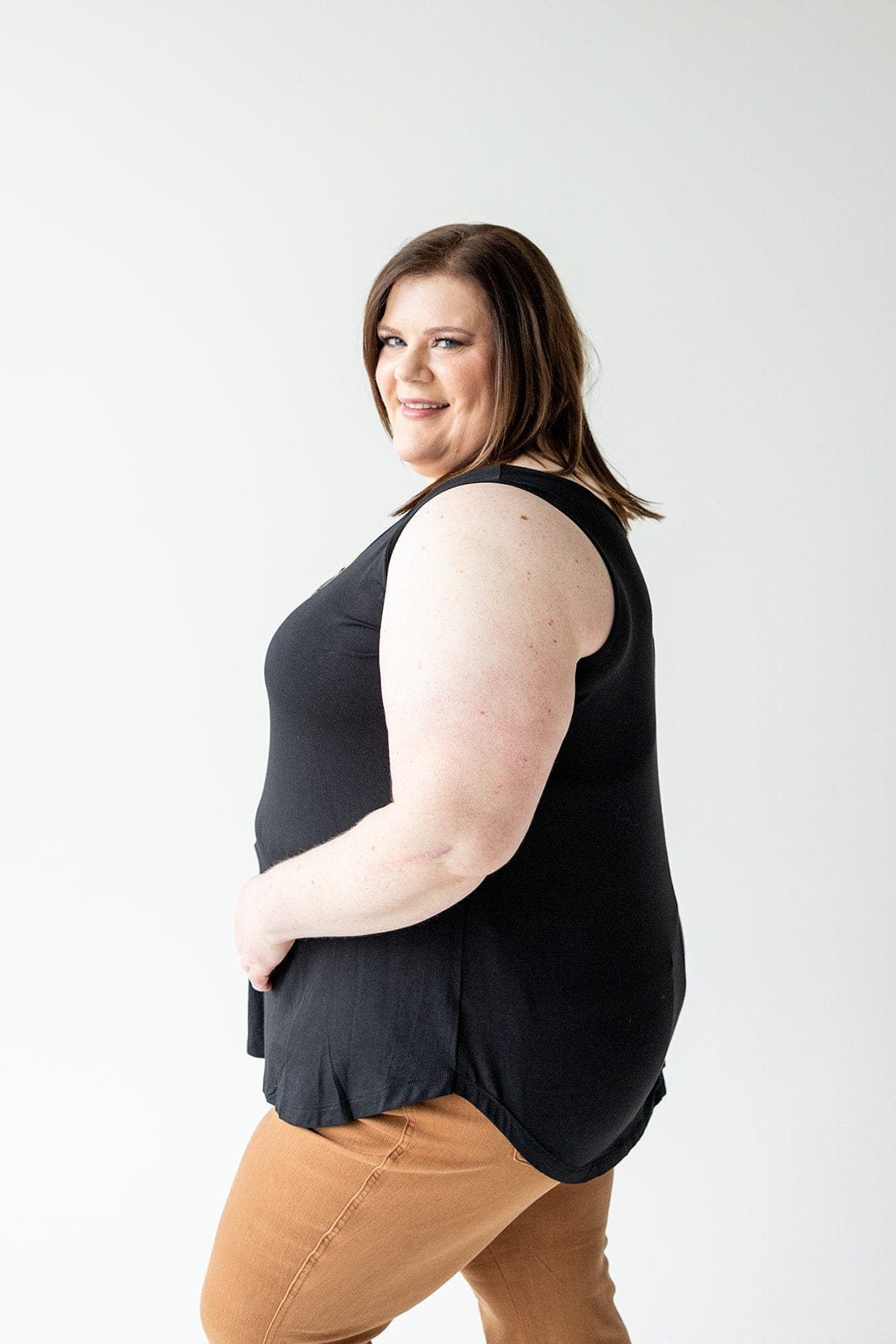 A woman with shoulder-length brown hair smiles while posing sideways. She is wearing a BASIC ROUND NECK TUNIC TANK IN BLACK by Love Marlow and brown pants, standing against a plain white background.