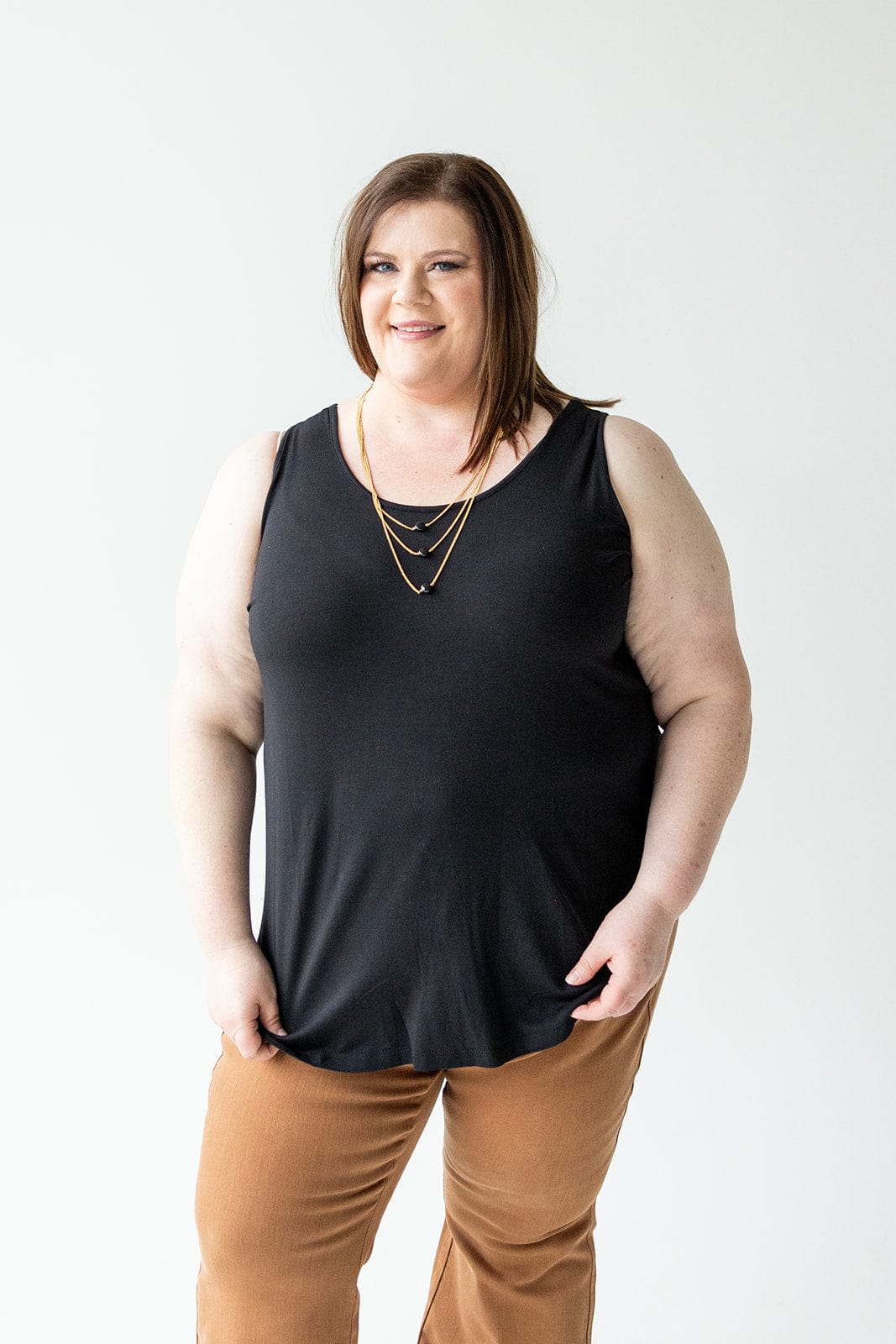 Person with brown hair, wearing a Love Marlow BASIC ROUND NECK TUNIC TANK IN BLACK featuring wide tank straps and a high-low rounded hem, along with brown pants. They are standing against a plain white background and smiling at the camera.