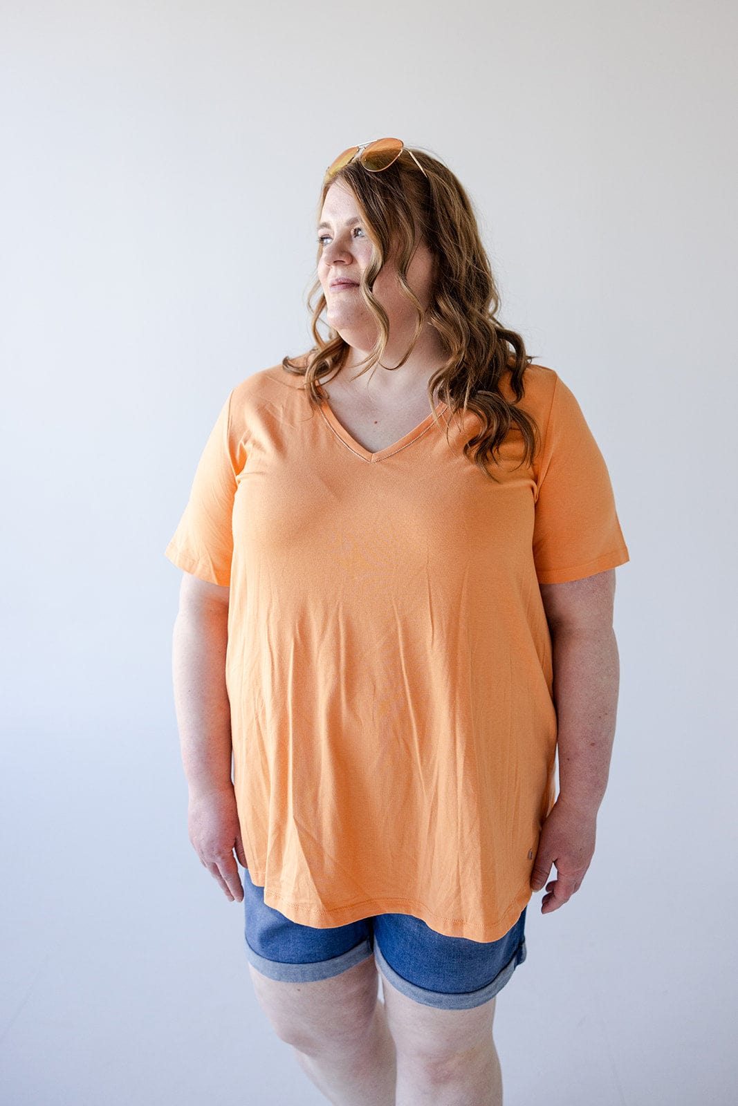 Person with long wavy hair wearing a BASIC V-NECK TEE WITH SPARKLE IN TANGERINE by Love Marlow, denim shorts, and sunglasses on their head, standing against a plain background.
