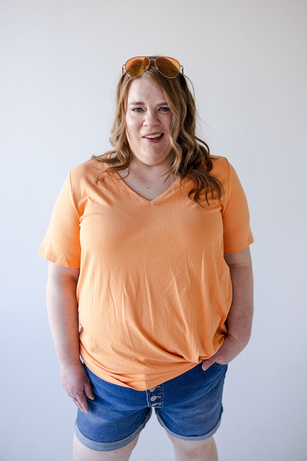 A woman with long hair wearing a BASIC V-NECK TEE WITH SPARKLE IN TANGERINE by Love Marlow, blue jean shorts, and sunglasses on her head stands against a plain white background.