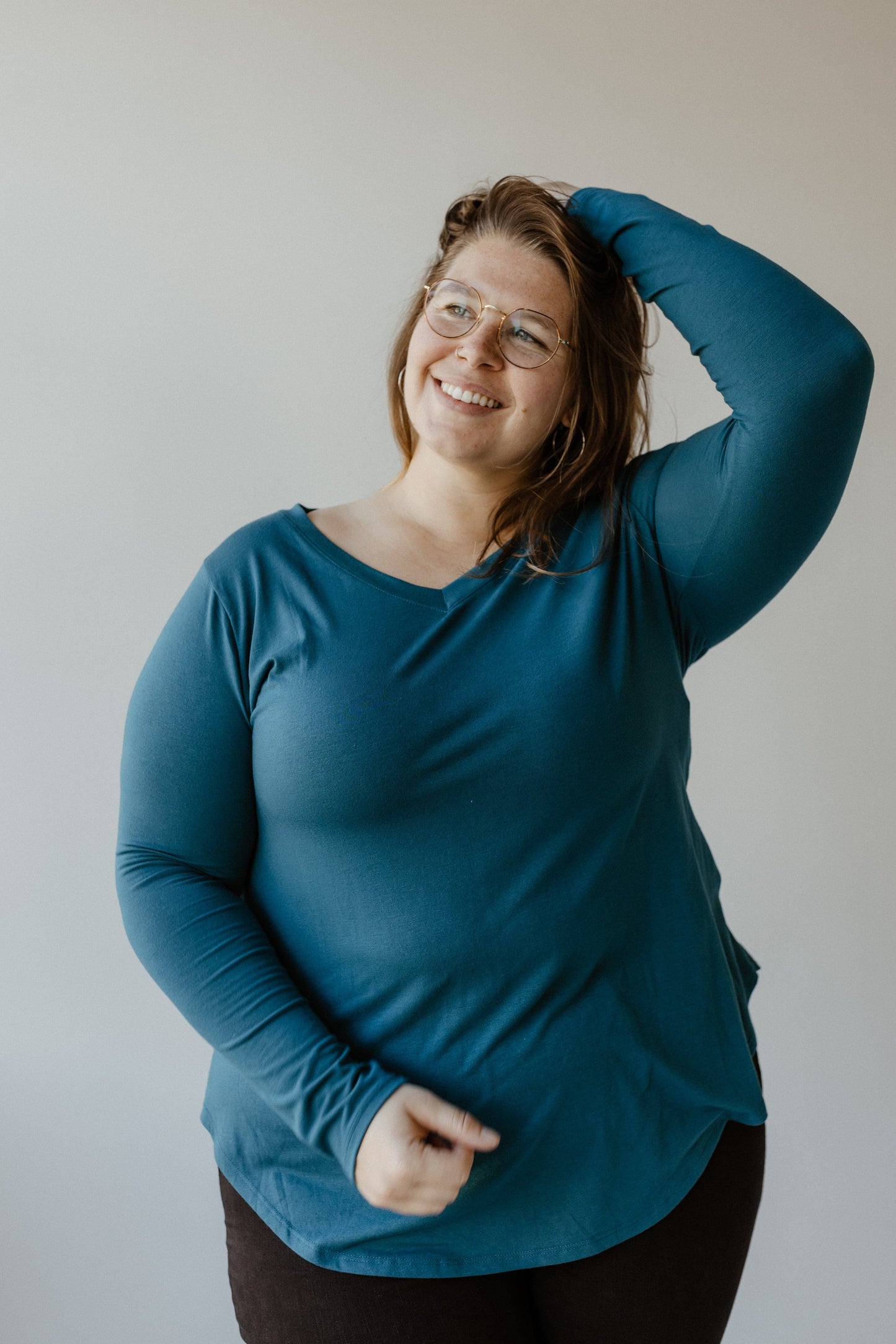 A person in glasses and a slim-fit teal long-sleeve shirt stands against a plain background, smiling with one hand on their head.