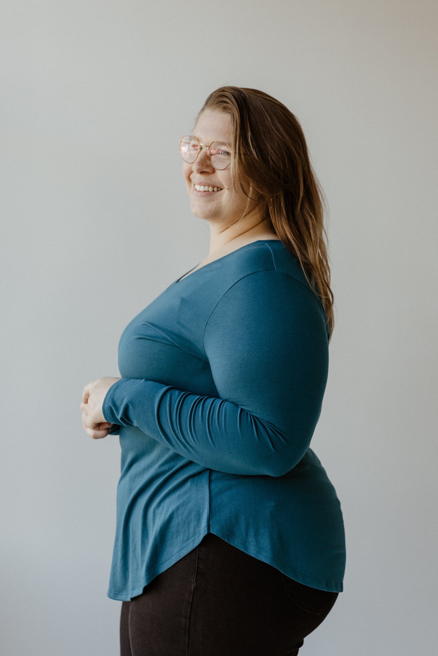 A person wearing glasses and a slim fit teal long-sleeve top is smiling and standing against a plain background.
