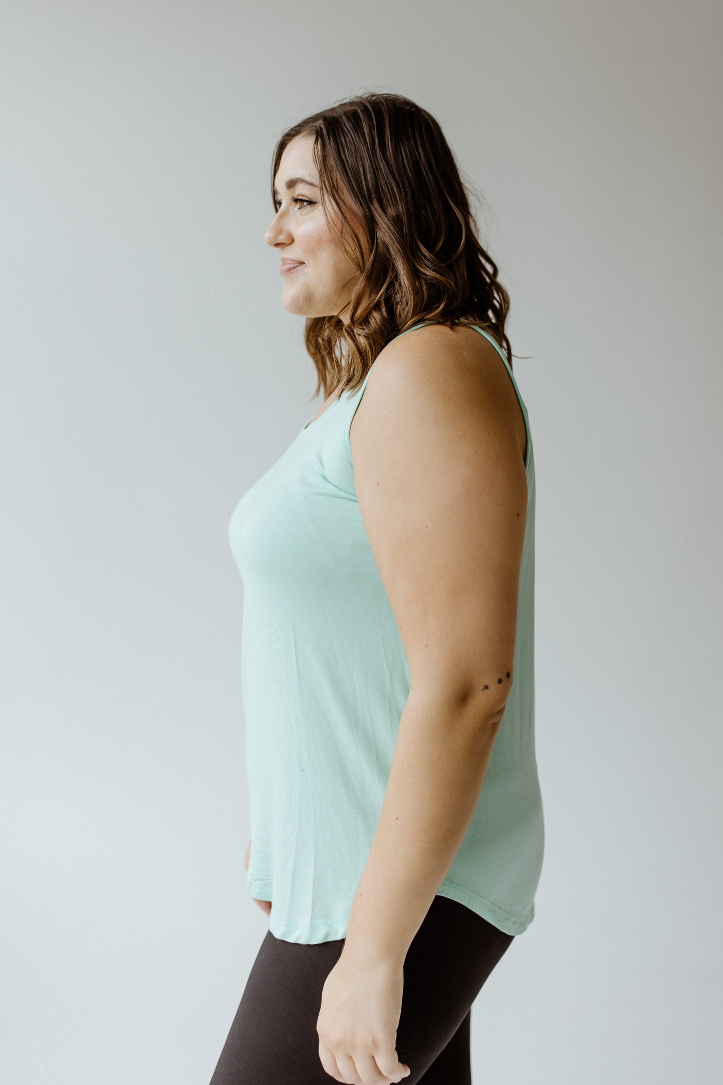 A person wearing a light green, plus-size basic round neck tunic tank and black leggings is standing in profile against a plain background.
