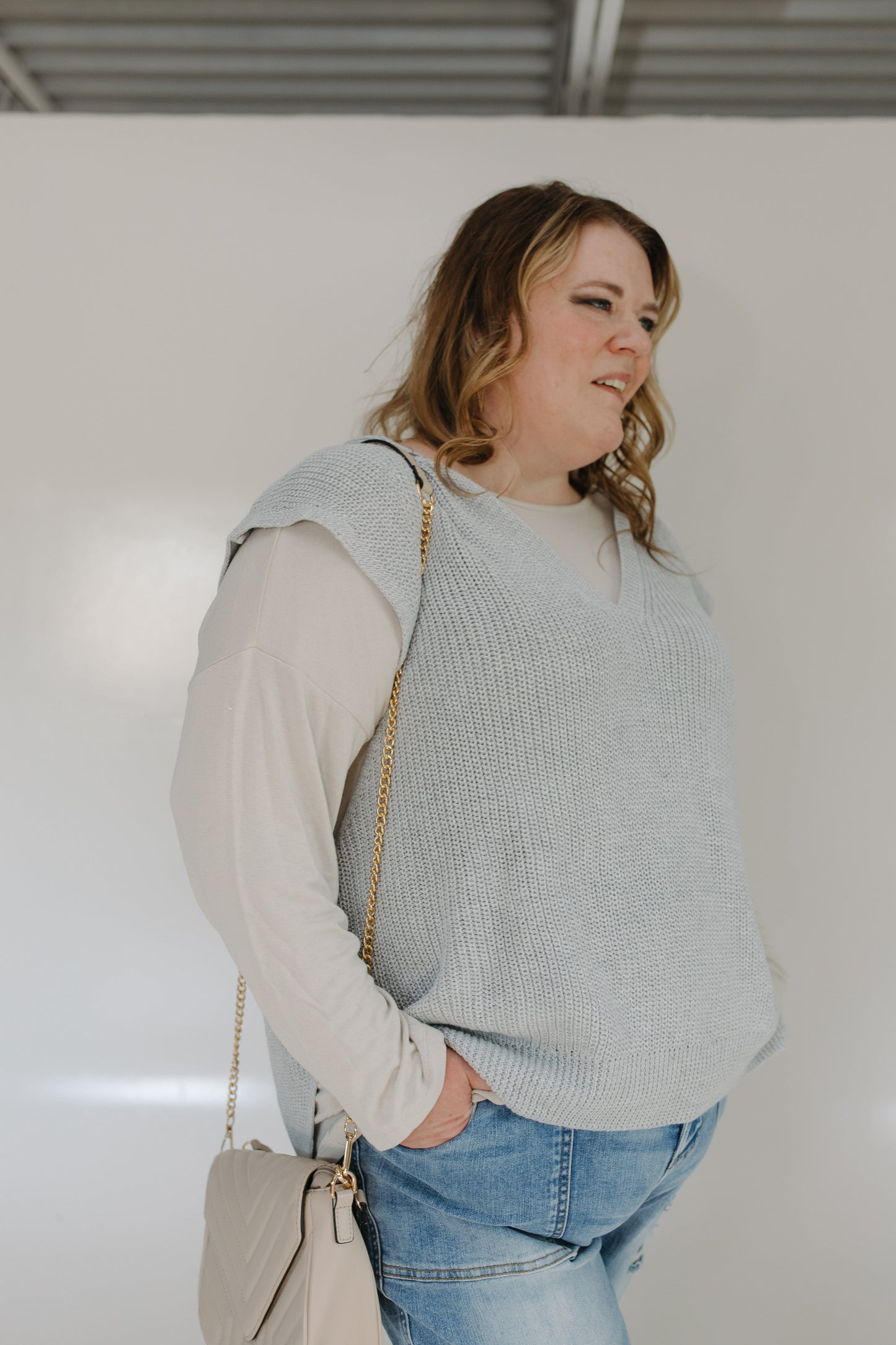 Woman wearing a light gray sweater vest over a beige shirt with blue jeans, standing against a plain white wall.