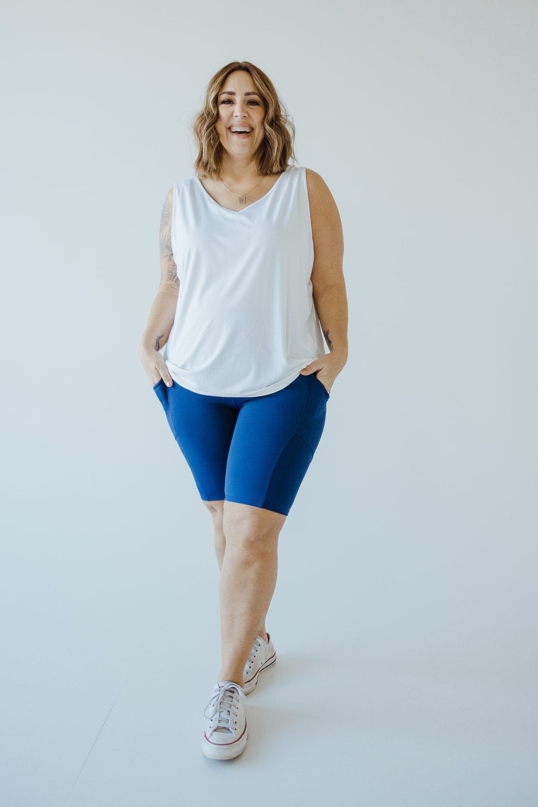 A woman in a white sleeveless top and the BIKER SHORTS WITH POCKETS IN MARINE by Love Marlow stands and smiles. She has her hands in her pockets and is wearing white sneakers.