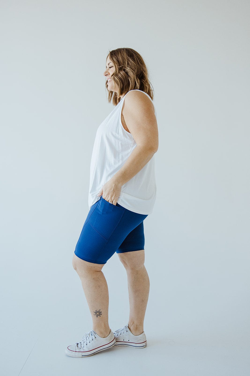 A person stands sideways, wearing a white sleeveless top, Love Marlow BIKER SHORTS WITH POCKETS IN MARINE featuring an athletic wide waistband, and white sneakers. A small star tattoo adorns their lower leg. The background is plain and light-colored.