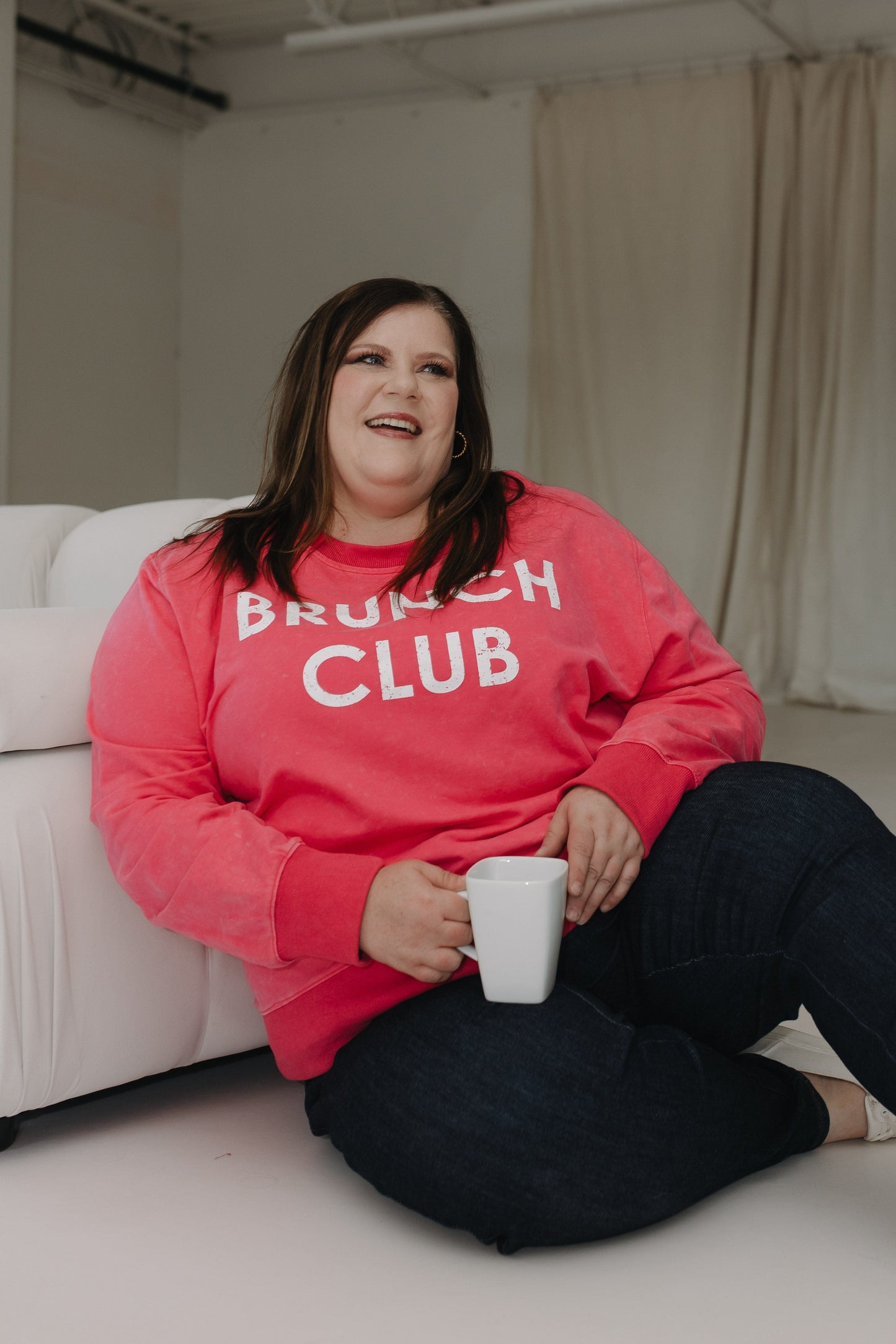 Woman wearing a 'Brunch Club' sweatshirt sitting on a couch holding a mug.