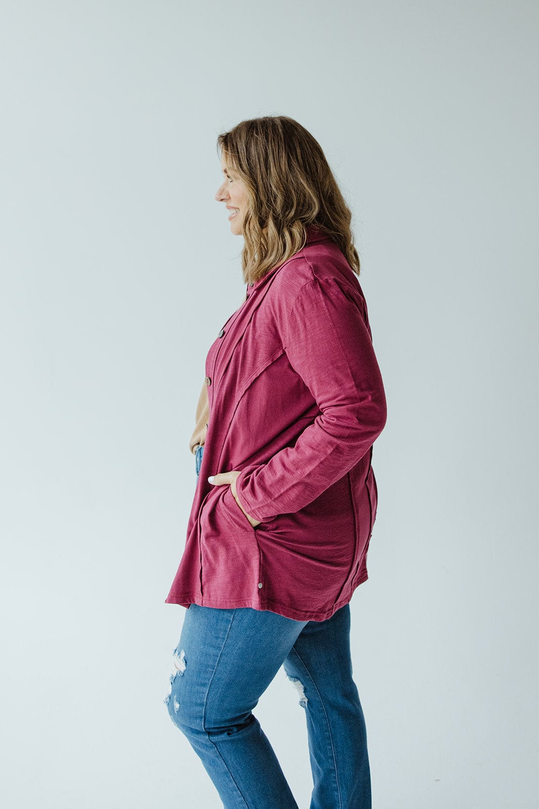 A woman wearing a cranberry button-up blouse with raw seam details from neon buddha and blue jeans stands in a light gray room, facing left with her hands in her pockets.
