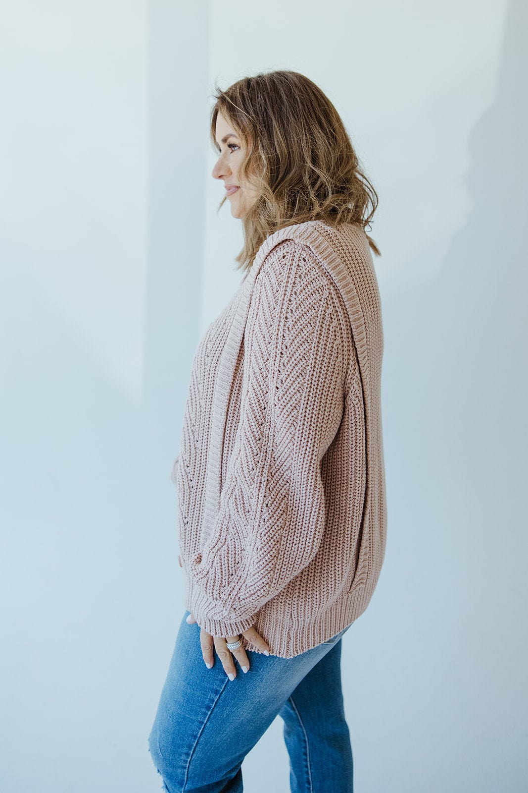 A person wearing a Yesta Button Front Cable Knit Cardigan in Grey Rose and blue jeans stands sideways against a plain white background.