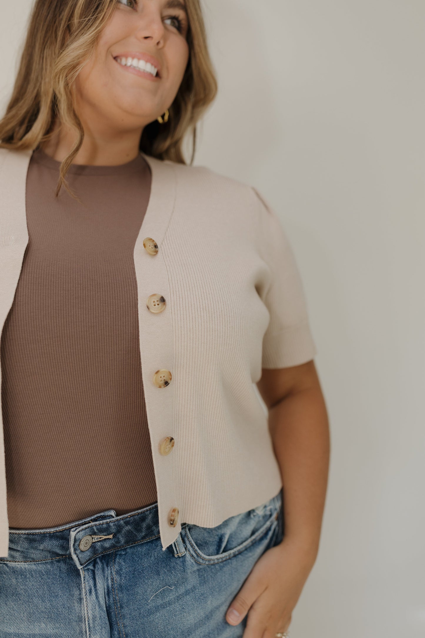 Woman wearing a beige cardigan over a brown top and blue jeans against a plain background