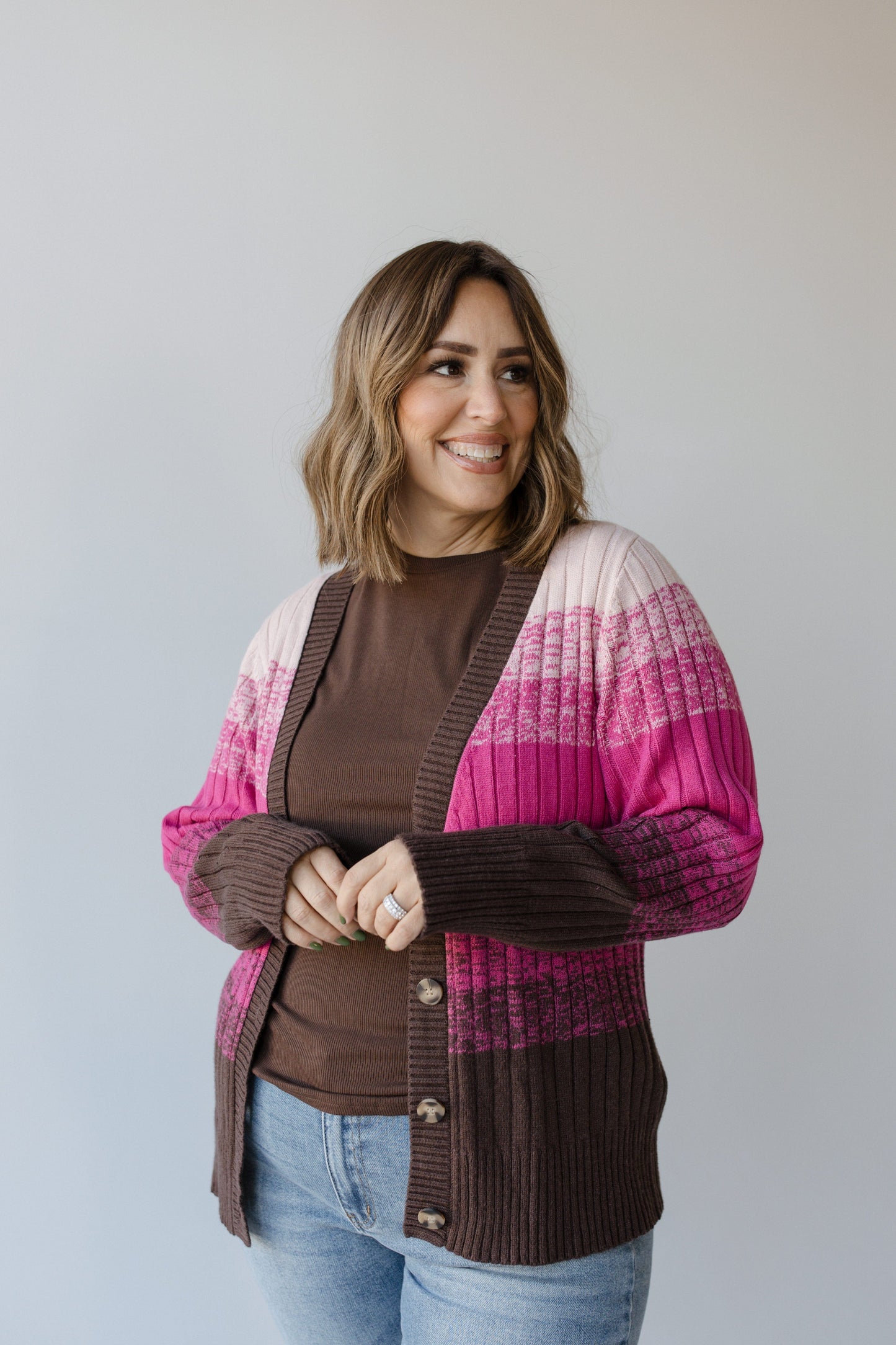 A person is smiling while wearing Yesta's CLASSIC CARDIGAN WITH COLOR FADE IN PINK and a pair of jeans, standing in front of a plain background.
