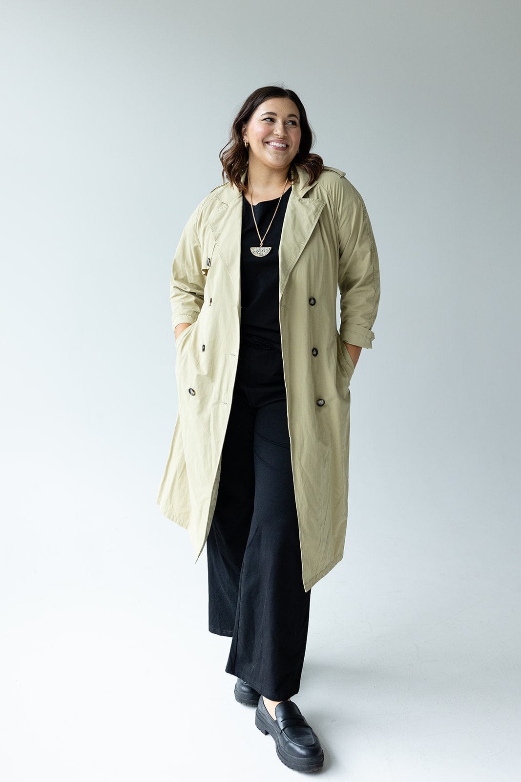 A person wearing the CLASSIC TRENCH COAT by Yesta, featuring double-breasted buttons in a classic beige color, stands with hands in pockets against a plain background, slightly smiling while dressed in a black outfit.