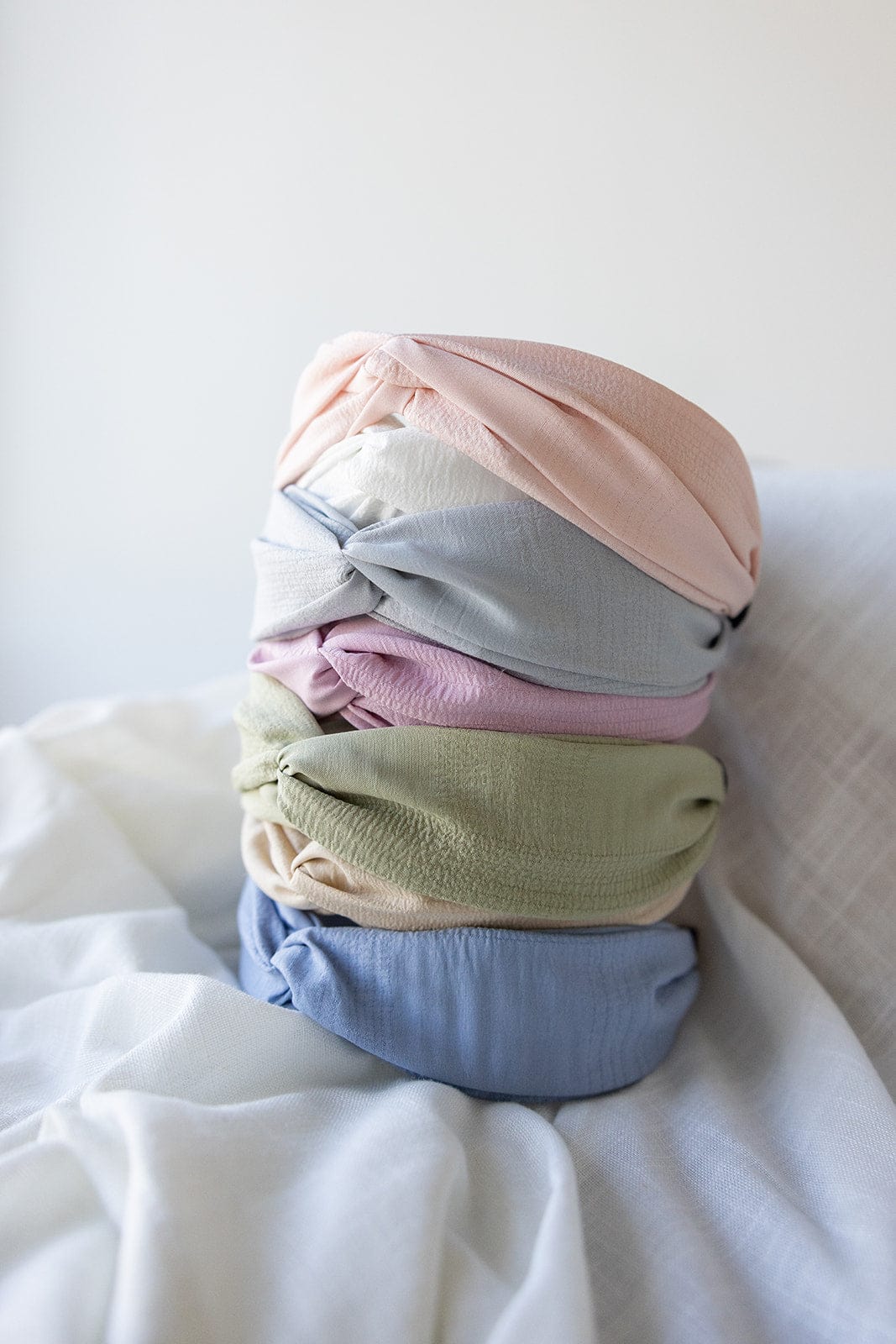 A stack of five CRINKLE TEXTURE HEADBAND WITH KNOT IN FRESH PINK by Love Marlow in pastel colors—pink, light blue, sage green, beige, and lavender—crafted from flexible material rests on a white cloth surface.