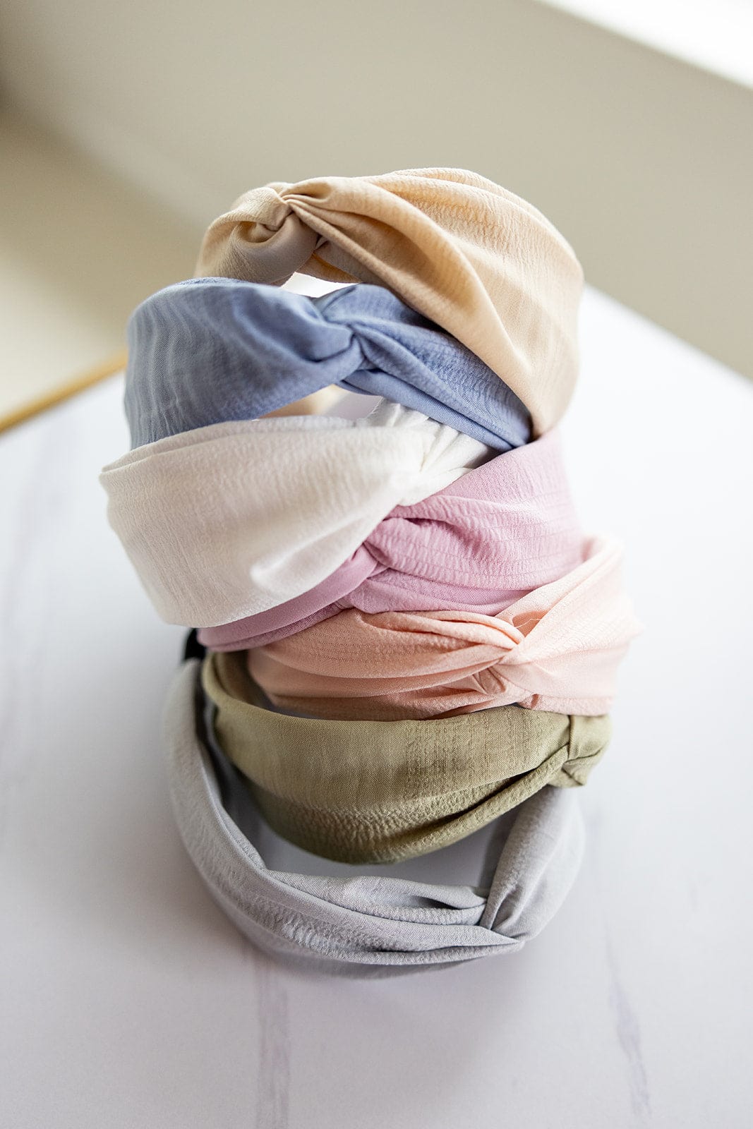 On a white surface lies a collection of six CRINKLE TEXTURE HEADBANDS WITH KNOT IN SAND by Love Marlow, displaying an arrangement of pastel colors with an elegant twisted knot design.