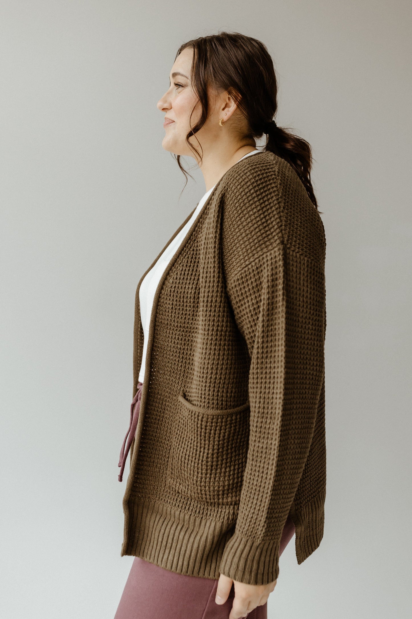 Woman in a crochet knit cardigan and maroon pants stands against a plain background, facing left.
