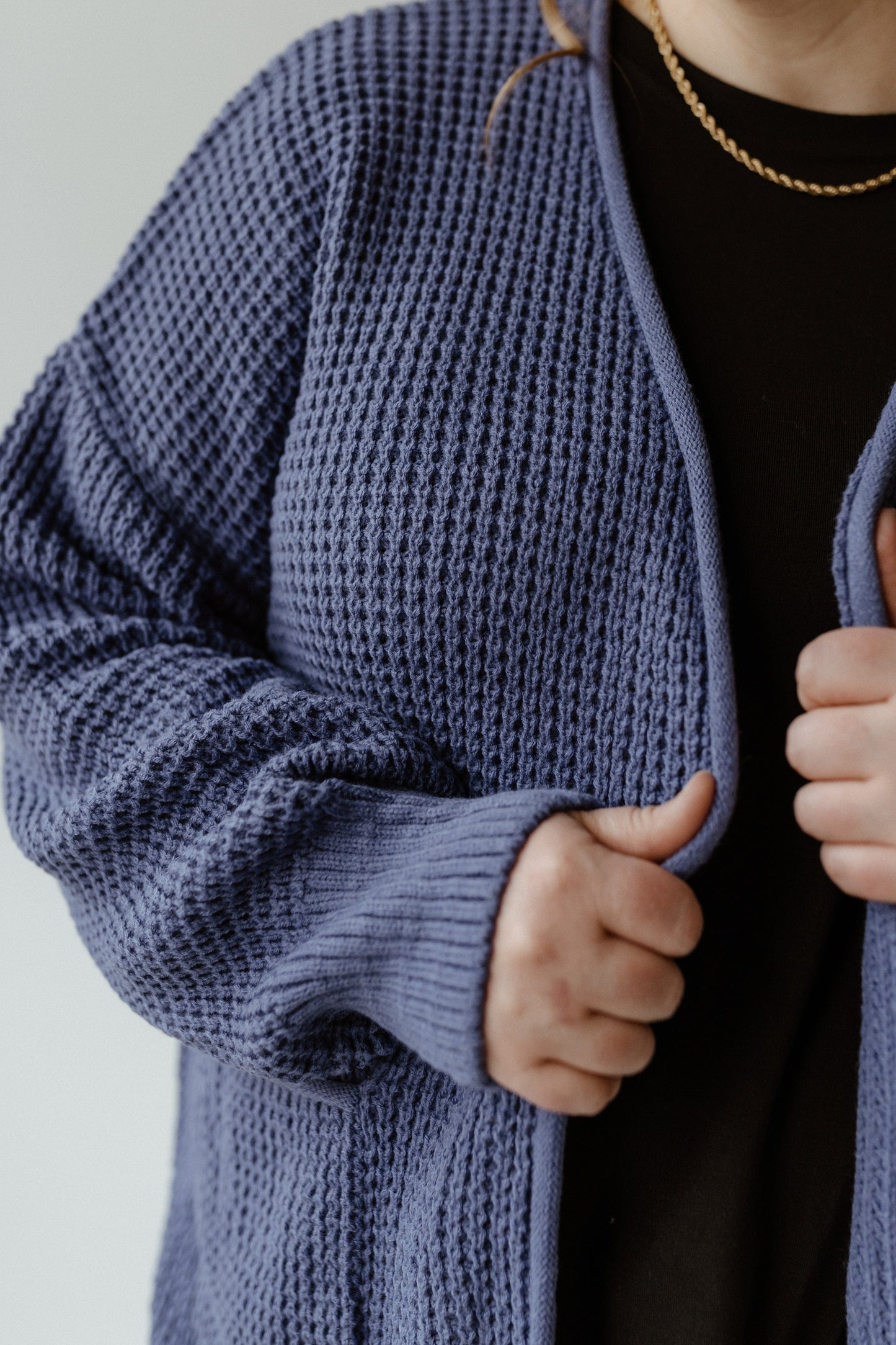 A person wearing a crochet knit cardigan in French blue over a black shirt, delicately holding the cardigan closed with both hands.