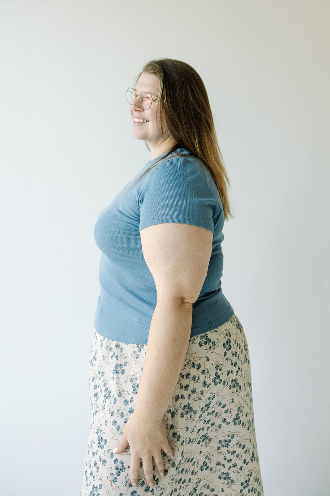 A person with glasses, wearing a Mikarose CROPPED SHORT SLEEVE RIBBED TEE IN ADRIFT and a floral skirt made from lightweight ribbed fabric, stands smiling against a plain white background.