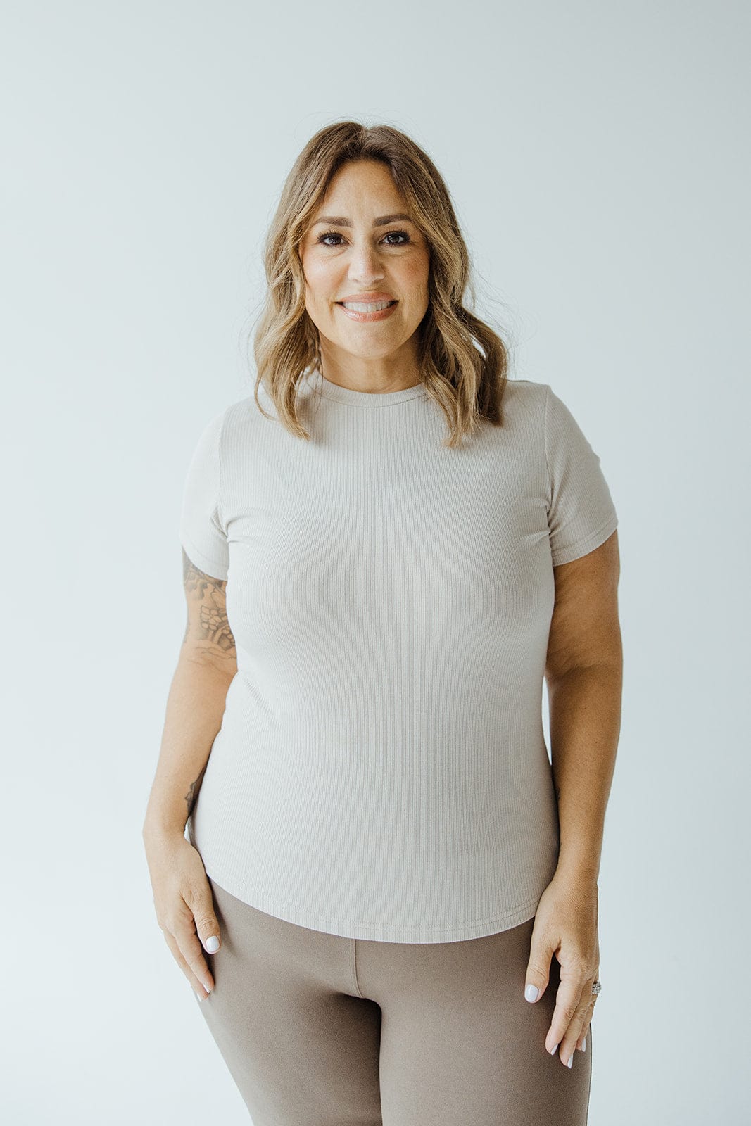 A person with shoulder-length hair, wearing a light beige shirt that's perfect for layering, and brown pants smiles at the camera against a plain background.