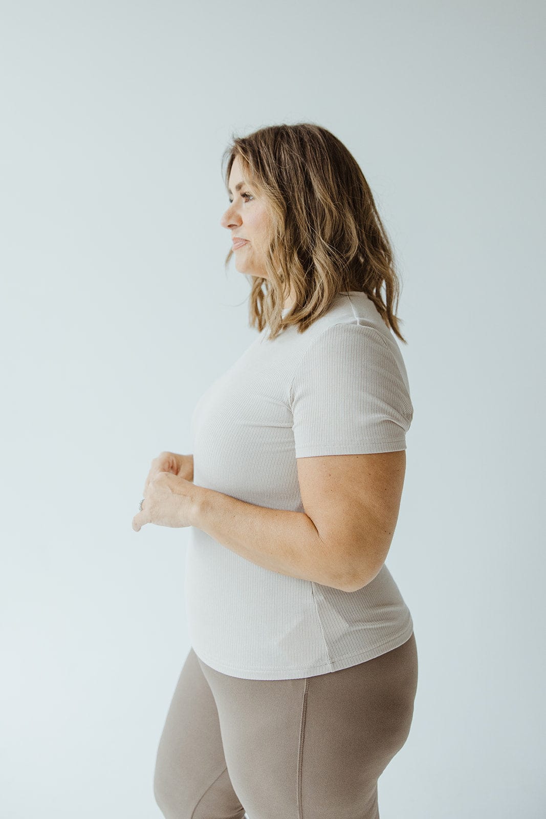A woman with shoulder-length hair stands in profile, wearing a white shirt and brown leggings that offer a lightweight feel, perfect for layering against the plain background.