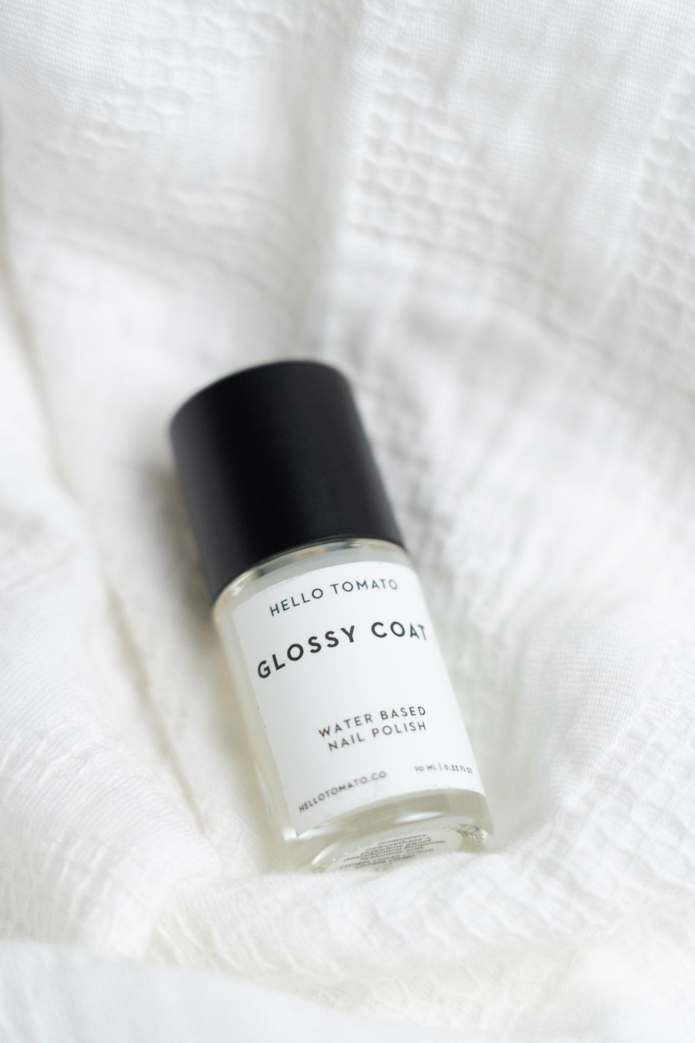 Bottle of 'Glossy Coat' nail polish by Hello Tomato on a white textured background