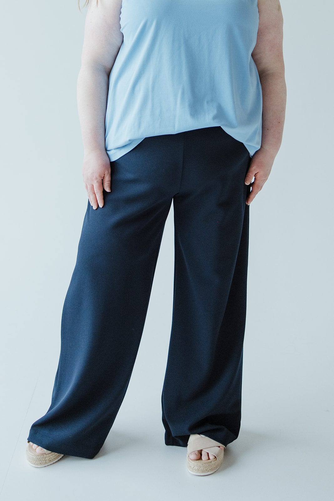 Person wearing a blue sleeveless top, dark wide-leg dress pants crafted from lightweight fabric, and sandals, standing in front of a plain white background.