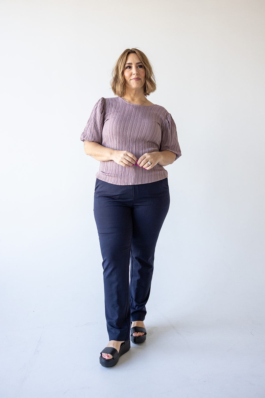 A woman with short hair stands against a plain white background, donning a mauve top and luxurious dress pants paired with black sandals.