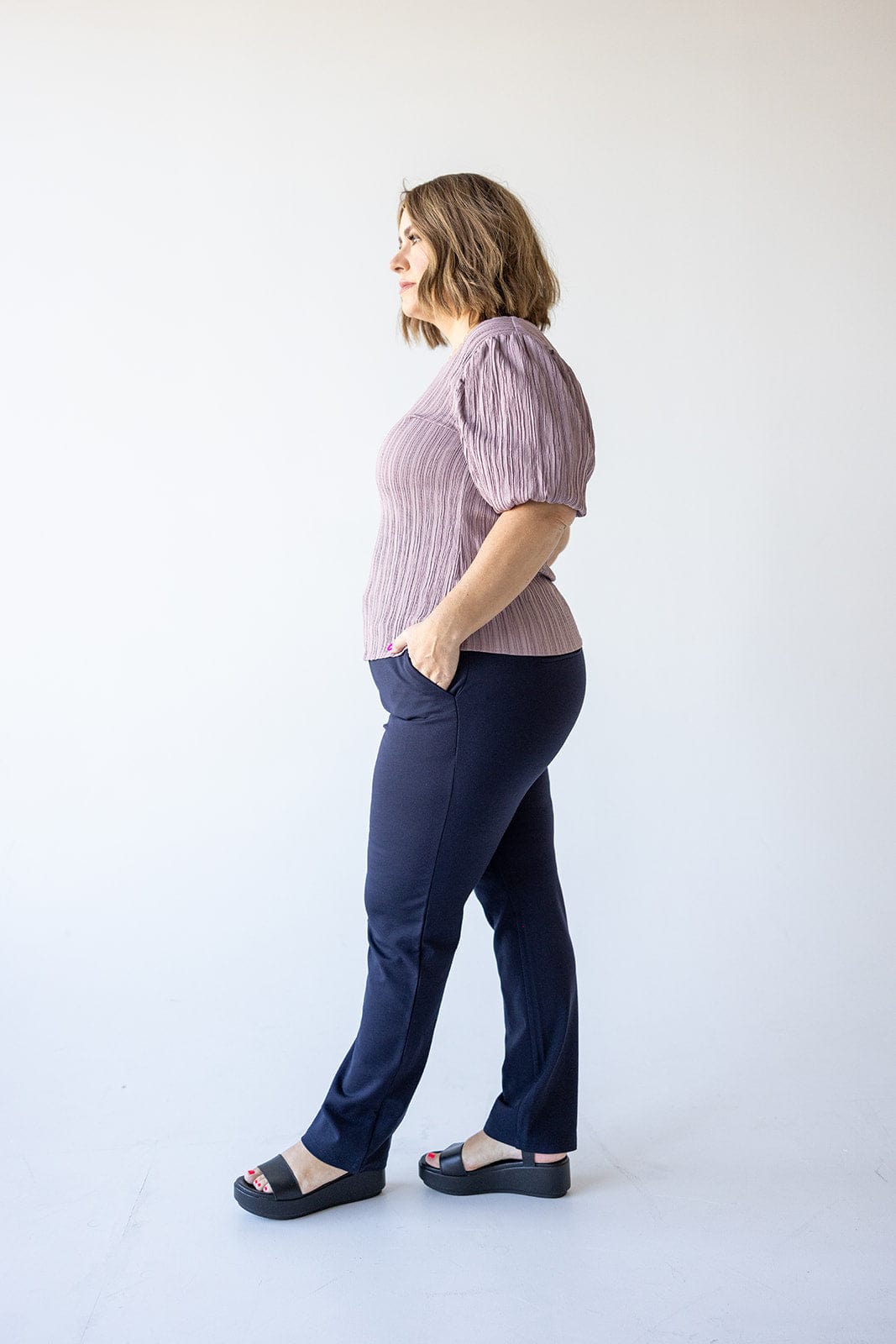 A person stands in profile, wearing a pink top and luxurious dress pants with hands in pockets, the smoothing technology of the fabric ensuring a sleek silhouette against a plain background.