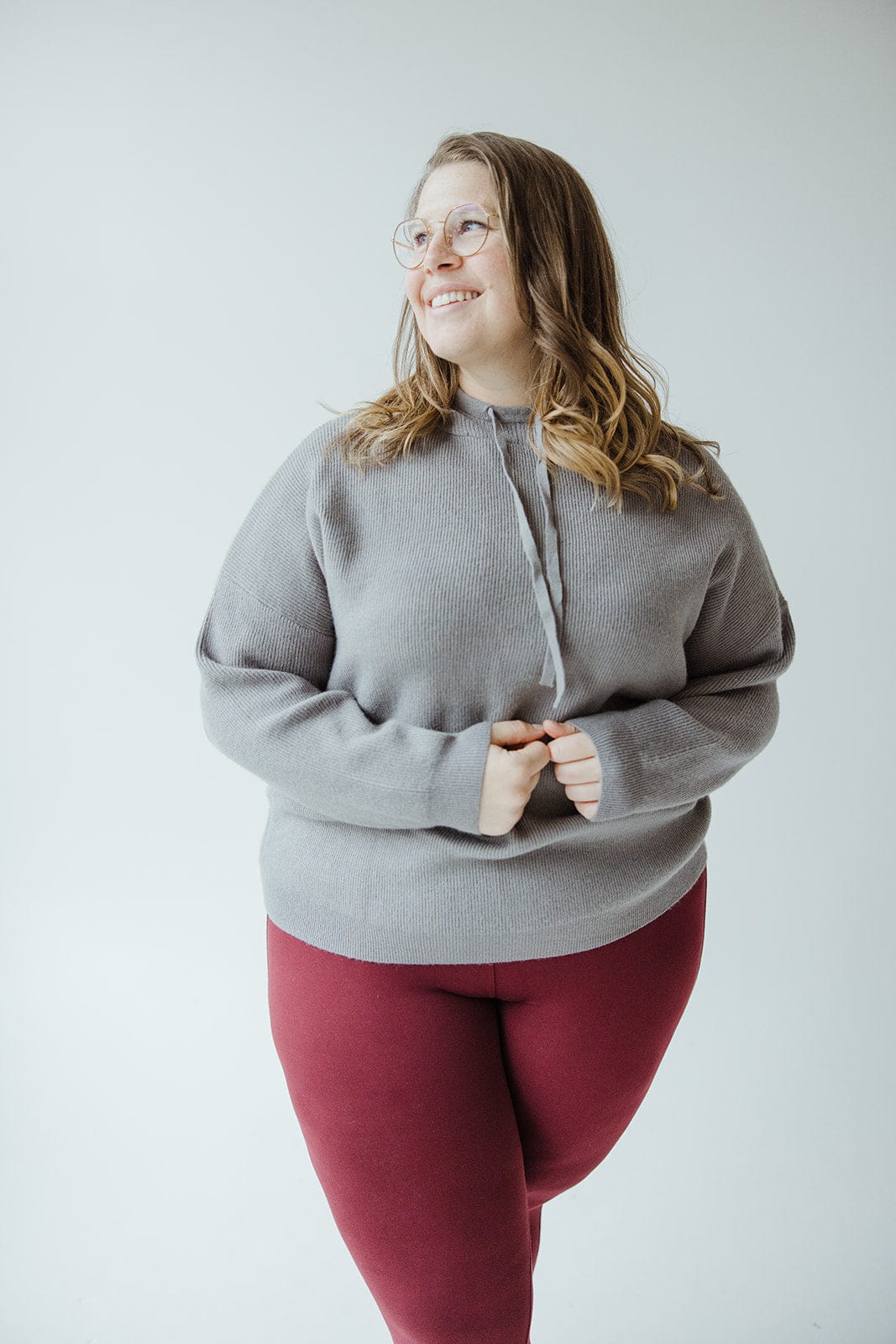 A person wearing glasses, a flattering silhouette created by a luxurious knit material in a gray sweater, and maroon leggings stands smiling against a plain white background.
