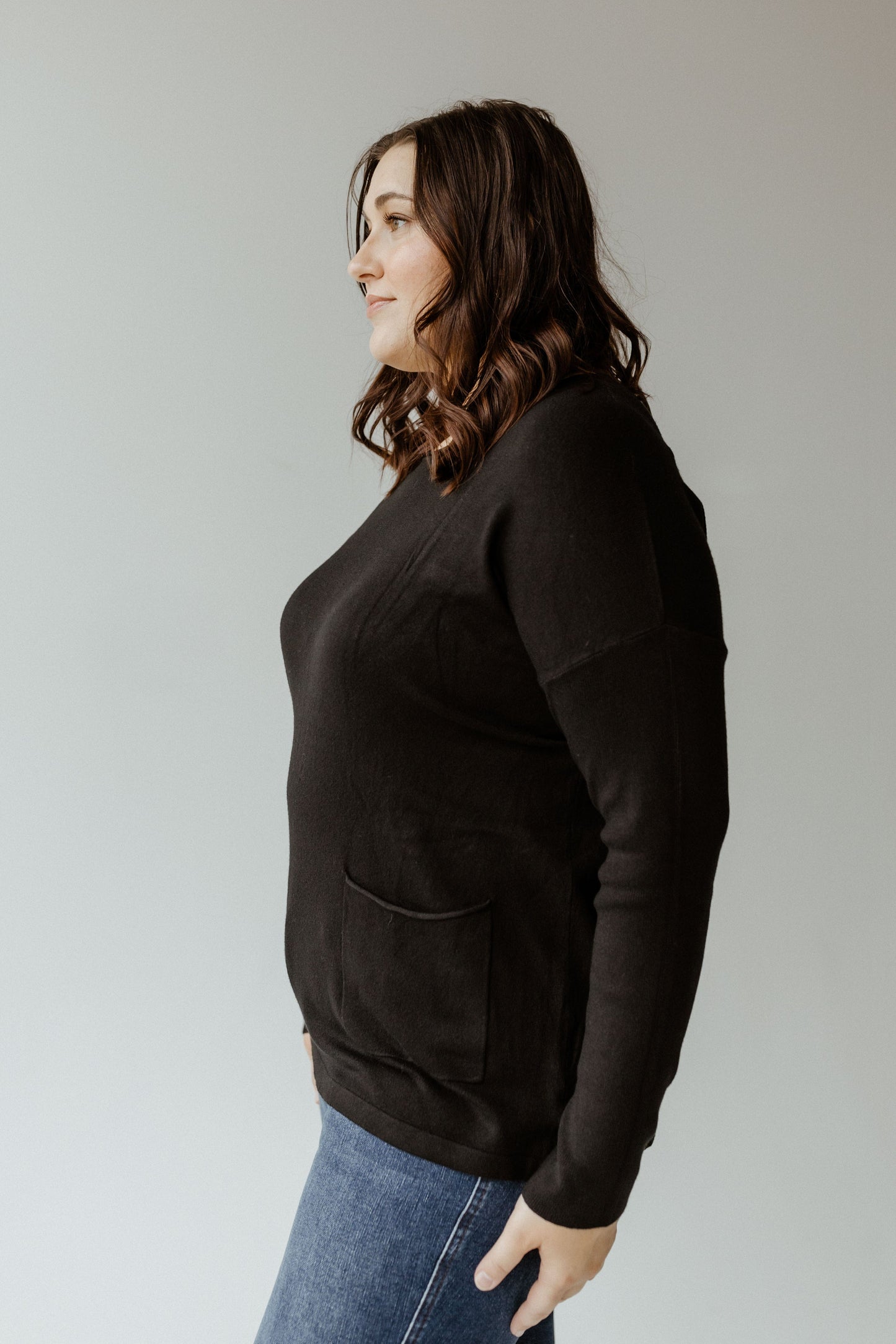 A person with shoulder-length brown hair is wearing a black Essential Crew Neck Sweater and blue jeans, standing sideways against a plain background.