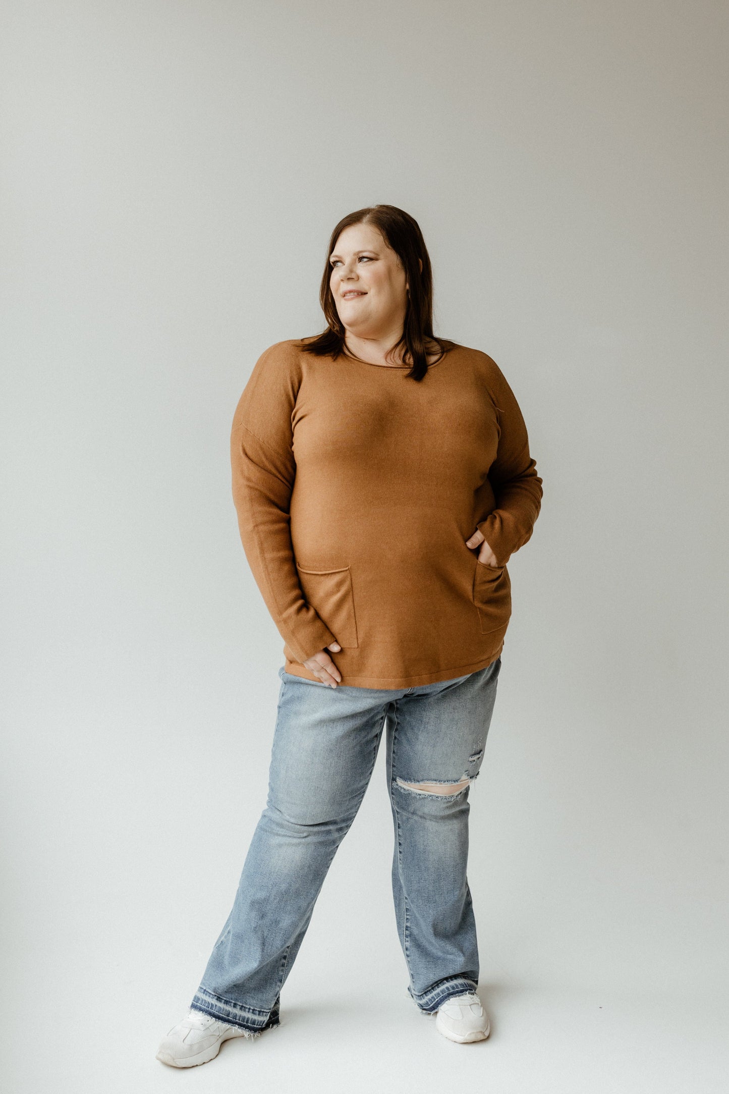 Person wearing an urban bronze crew neck sweater and ripped jeans, standing with hands in pockets against a neutral background.