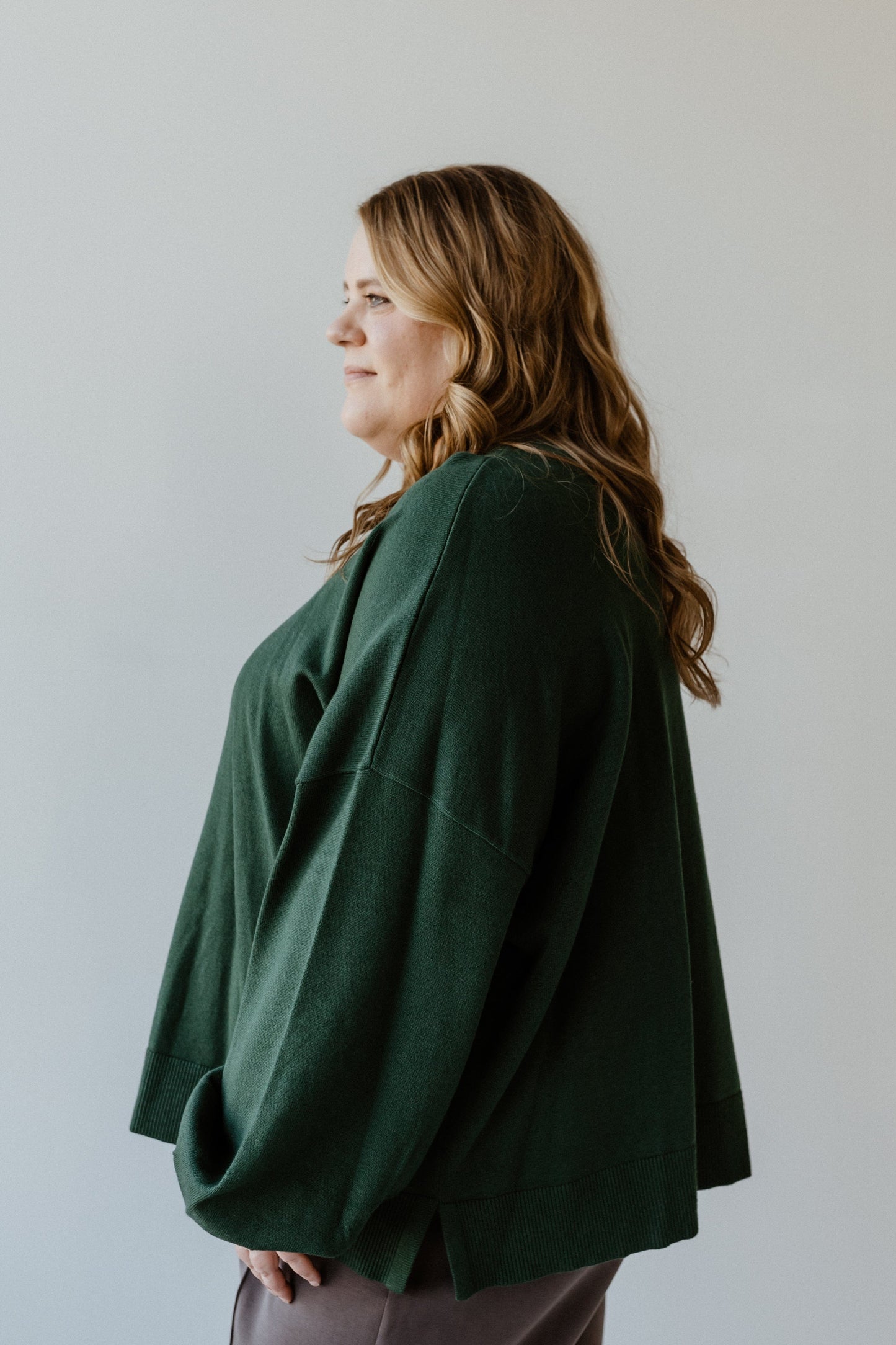 A woman with long hair wears an Essential Sweater, colored like a cut emerald, standing against a plain background, facing left.