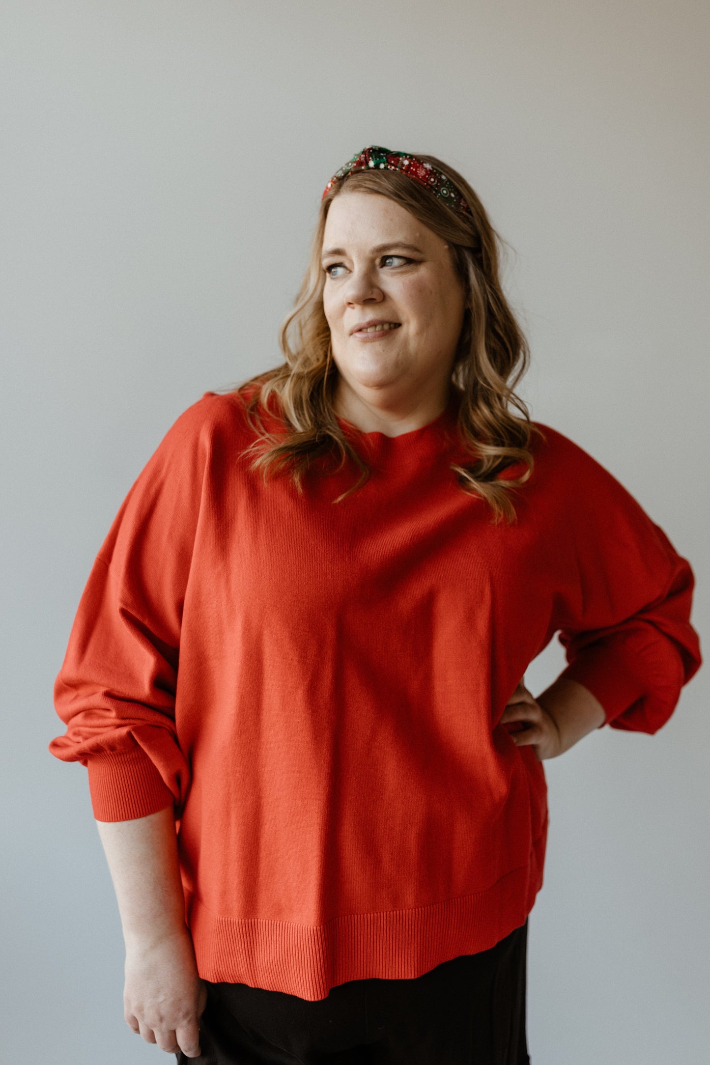 A person wearing a bold red sweater and jeweled headband stands against a plain background with a neutral expression.