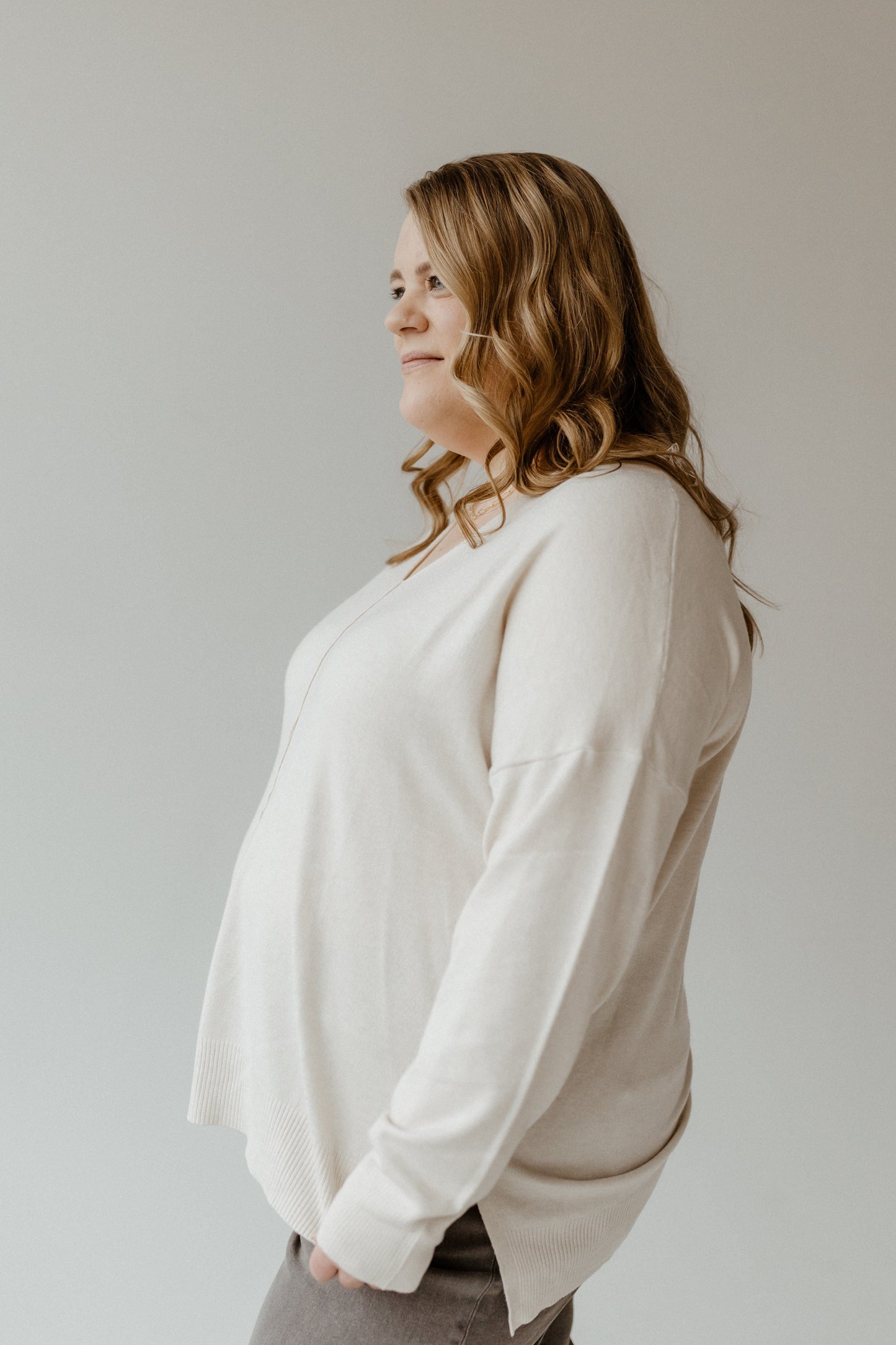 Side profile of a person with long wavy hair, wearing a slim fit light sweater with a front seam and pants, standing against a plain background.