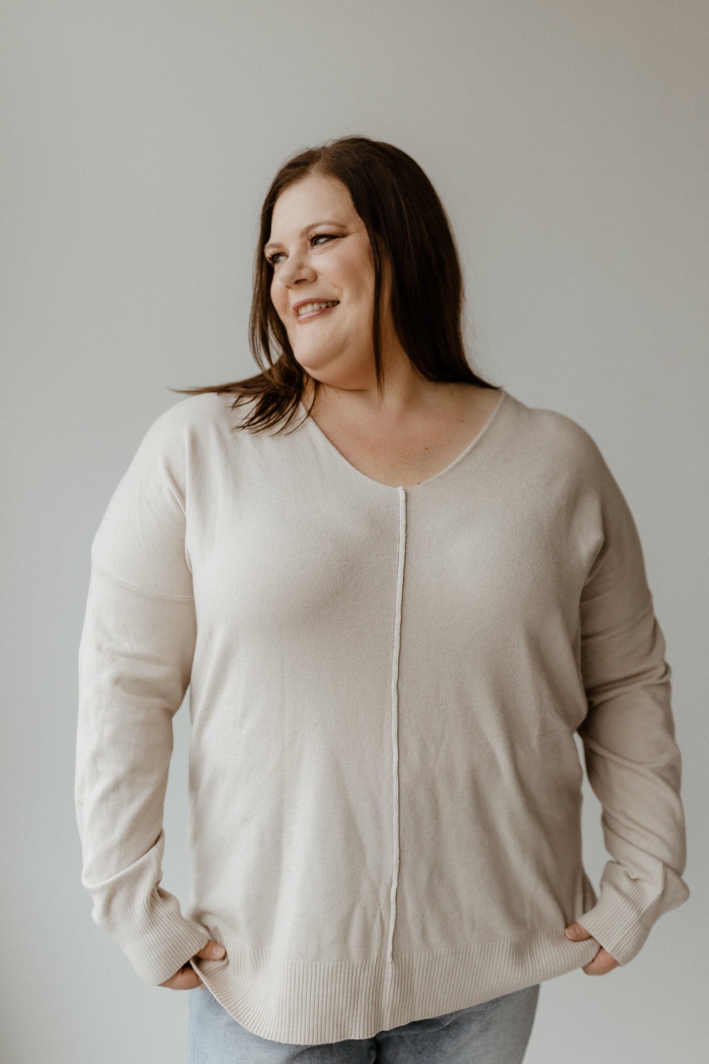 A person with long hair wearing a cozy, plus-size sweater and jeans smiles while looking to the side against a plain background.
