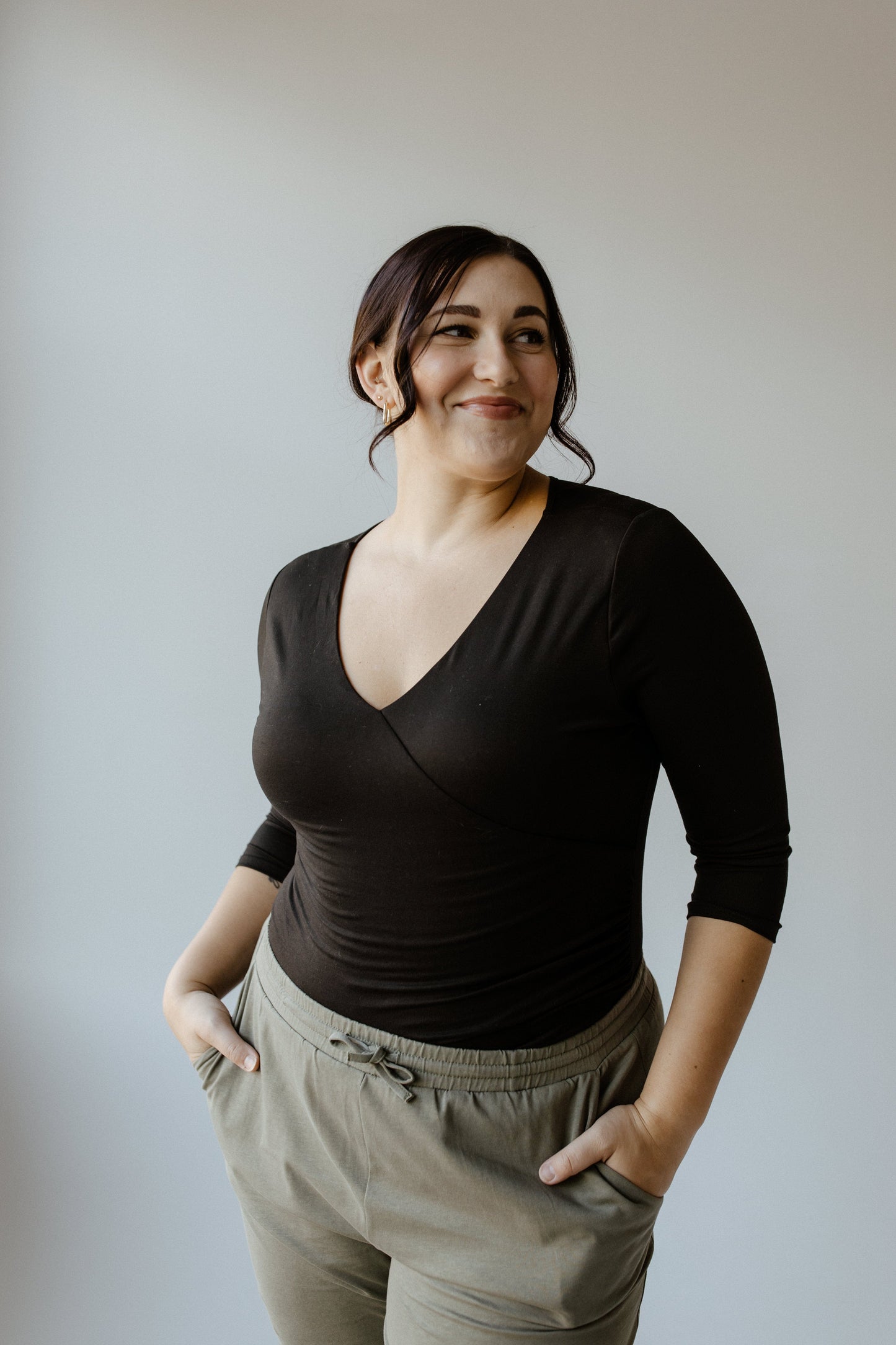 A person in a classic black sleek knit top and olive green pants stands smiling with hands in pockets against a light background.