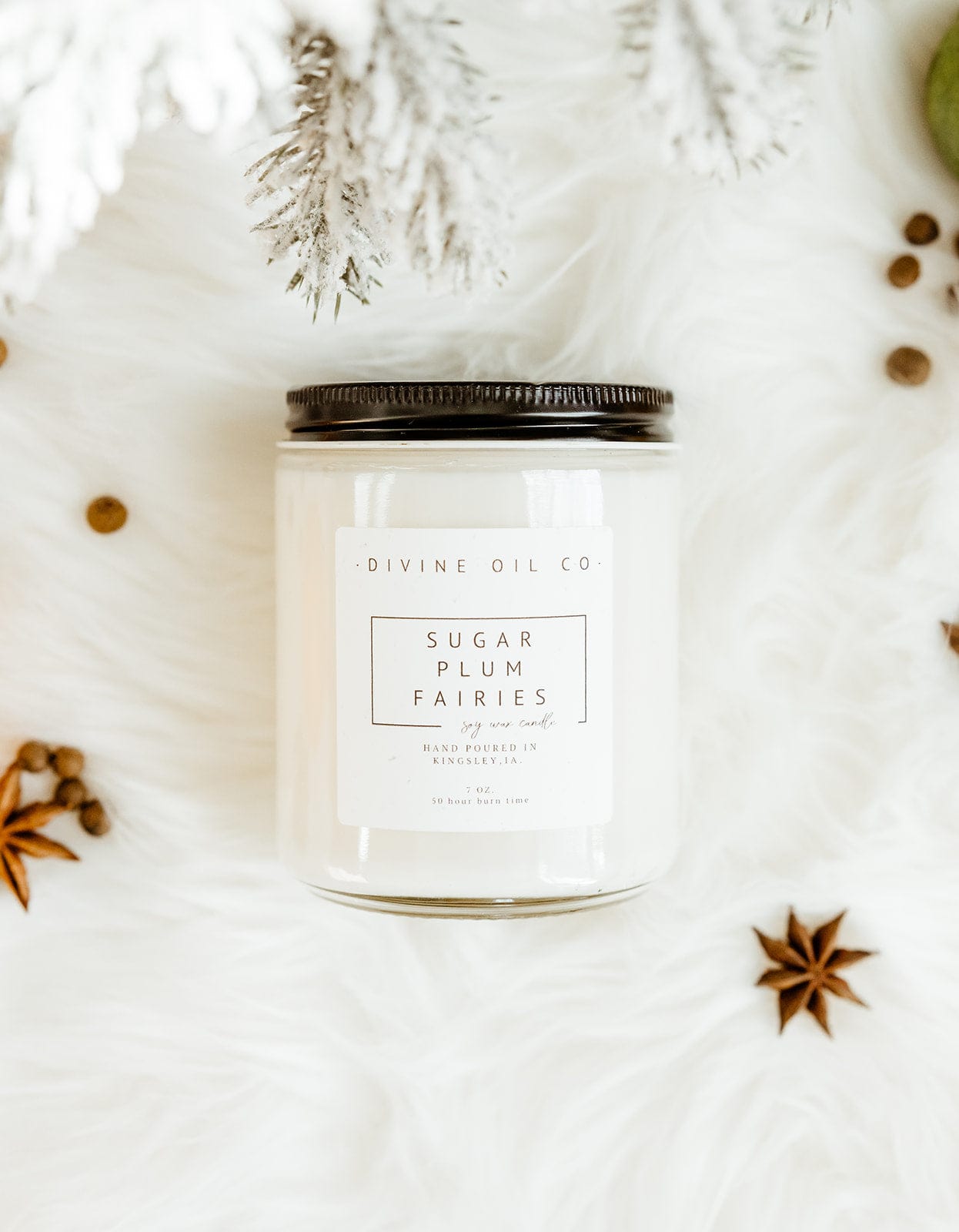 A jar candle labeled "Love Marlow HANDCRAFTED SOY WAX CANDLES IN SUGAR PLUM FAIRIES," crafted from Handcrafted Soy Wax Candles and phthalate-free fragrances, is placed on a white, fluffy surface surrounded by star anise and other small botanical elements.