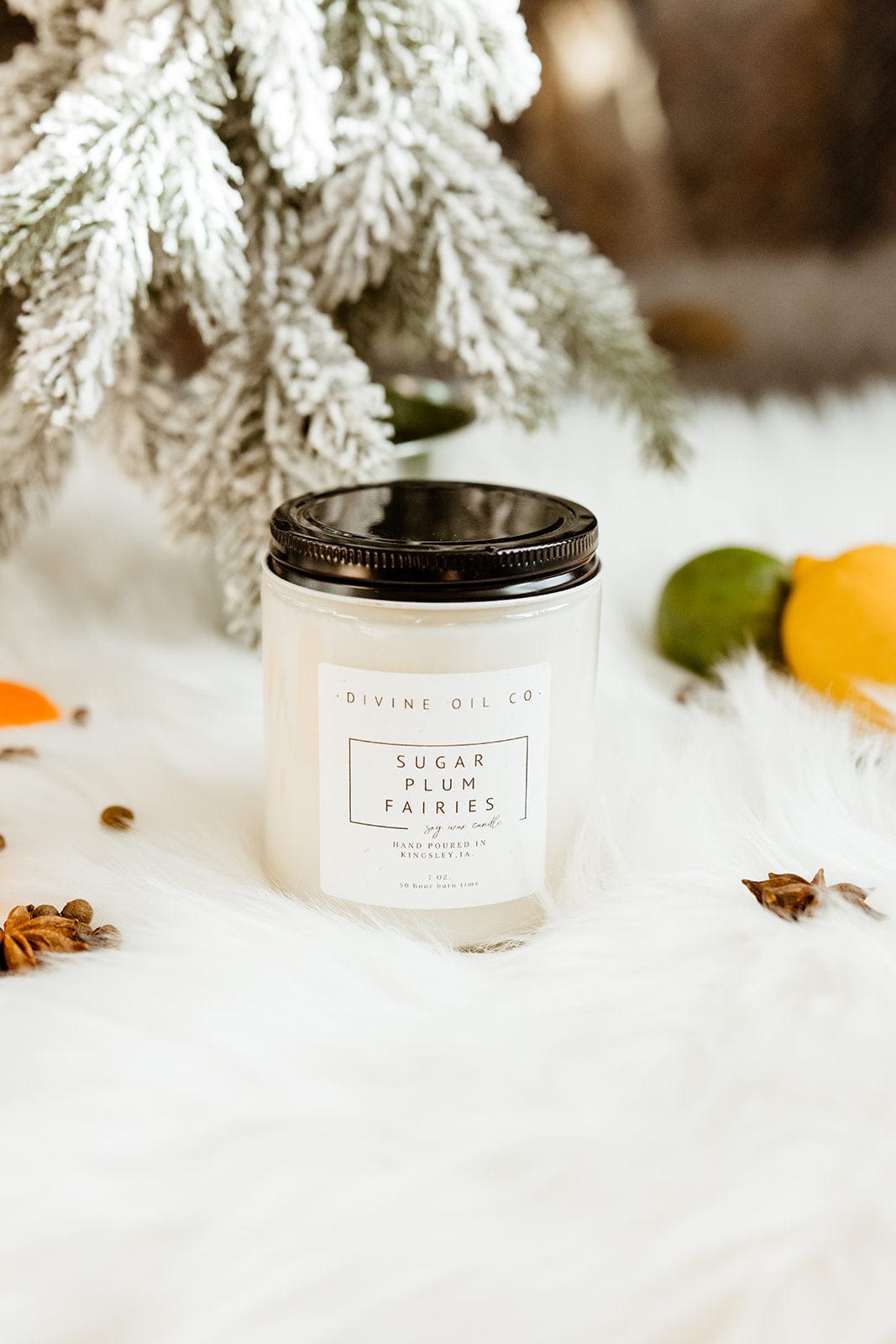 A white candle in a jar labeled "Love Marlow, HANDCRAFTED SOY WAX CANDLES IN SUGAR PLUM FAIRIES" sits on a fluffy white surface with a snow-covered branch and scattered spices in the background. This handcrafted soy wax candle, infused with phthalate-free fragrances, radiates an aura of seasonal charm.