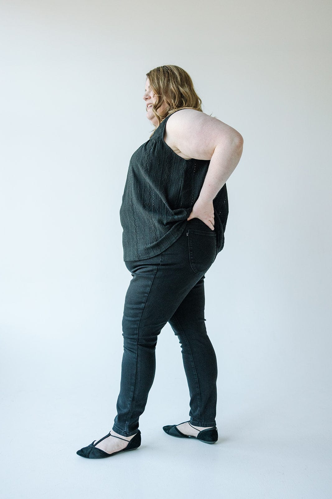 A person wearing a sleeveless black top, Judy Blue High Waist Pull-On Skinny Jeans in Black, and black shoes stands with a hand on their hip in a minimalist setting.