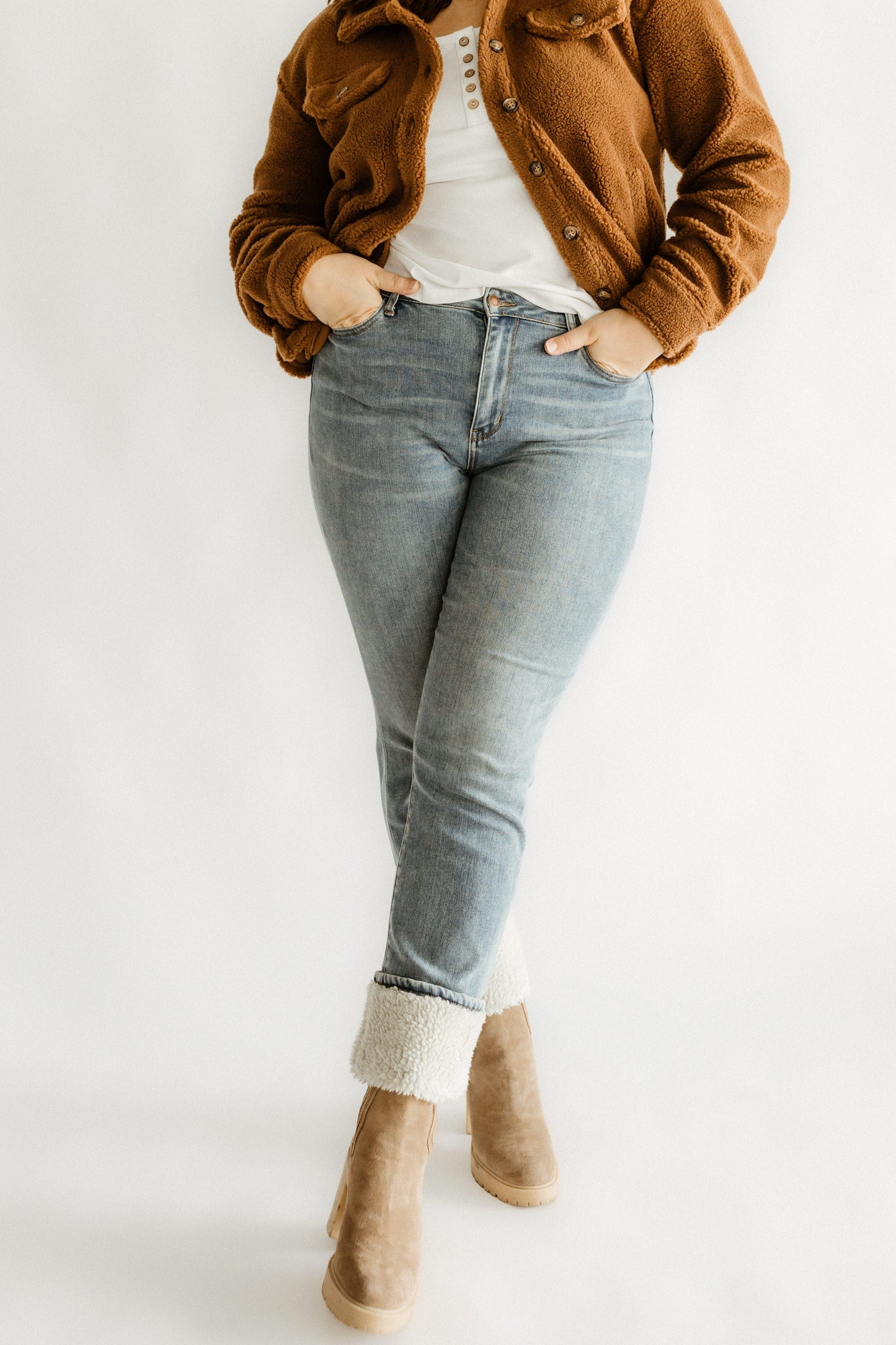 A person wearing a brown jacket, white shirt, medium wash jeans rolled up at the ankles, and tan boots stands against a plain background.