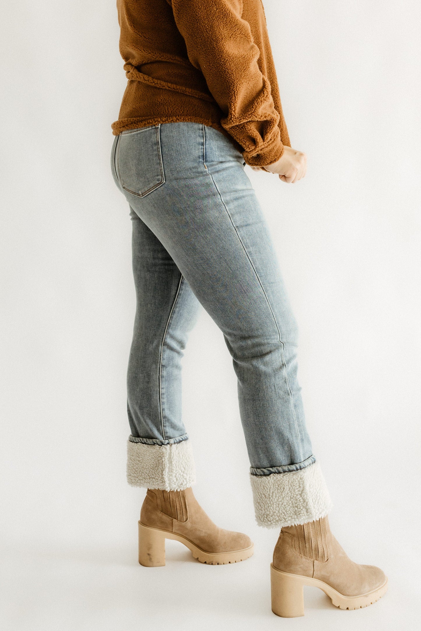 A person wearing high waist straight leg jeans, a brown sweater, and tan heeled boots stands against a plain background.