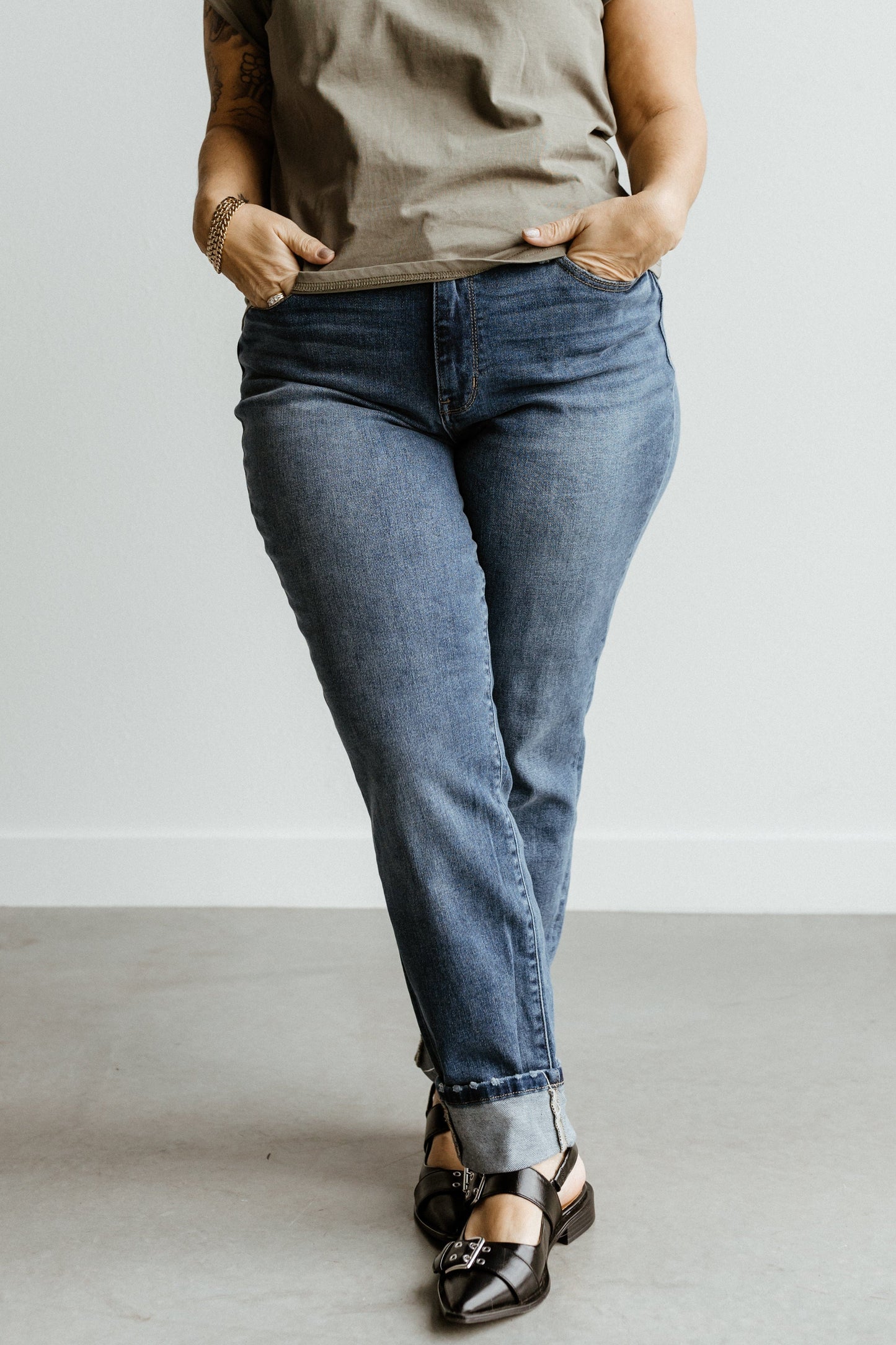 A person wearing a gray shirt and dark wash Judy Blue jeans with hands in pockets, standing on a sleek gray floor.