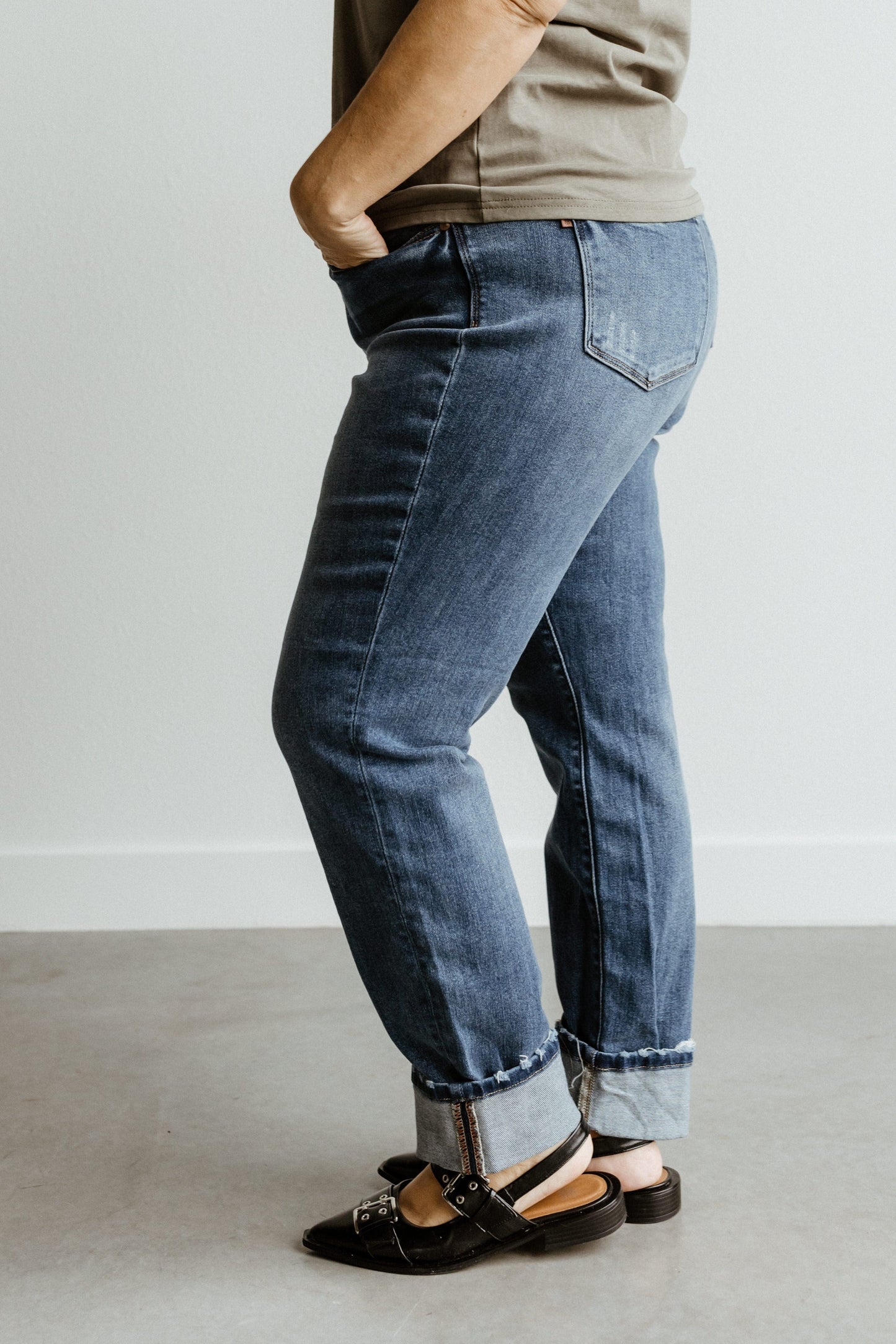 A person in dark wash Judy Blue mid-rise boyfriend jeans, hands in pockets, wearing black shoes, stands against a white wall on a gray floor.