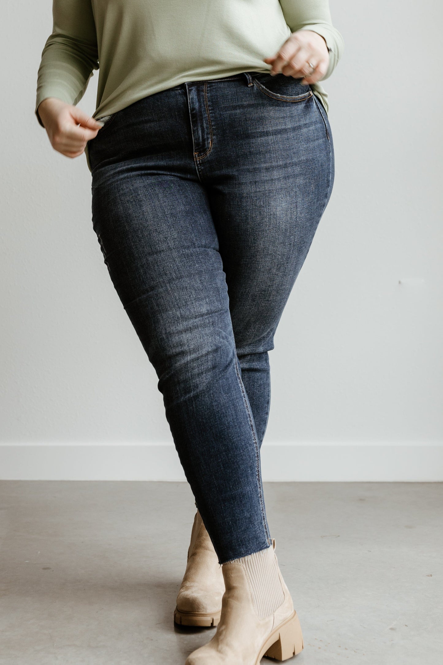 A person in a light green sweater and Judy Blue jeans stands on a light gray floor, wearing beige ankle boots.