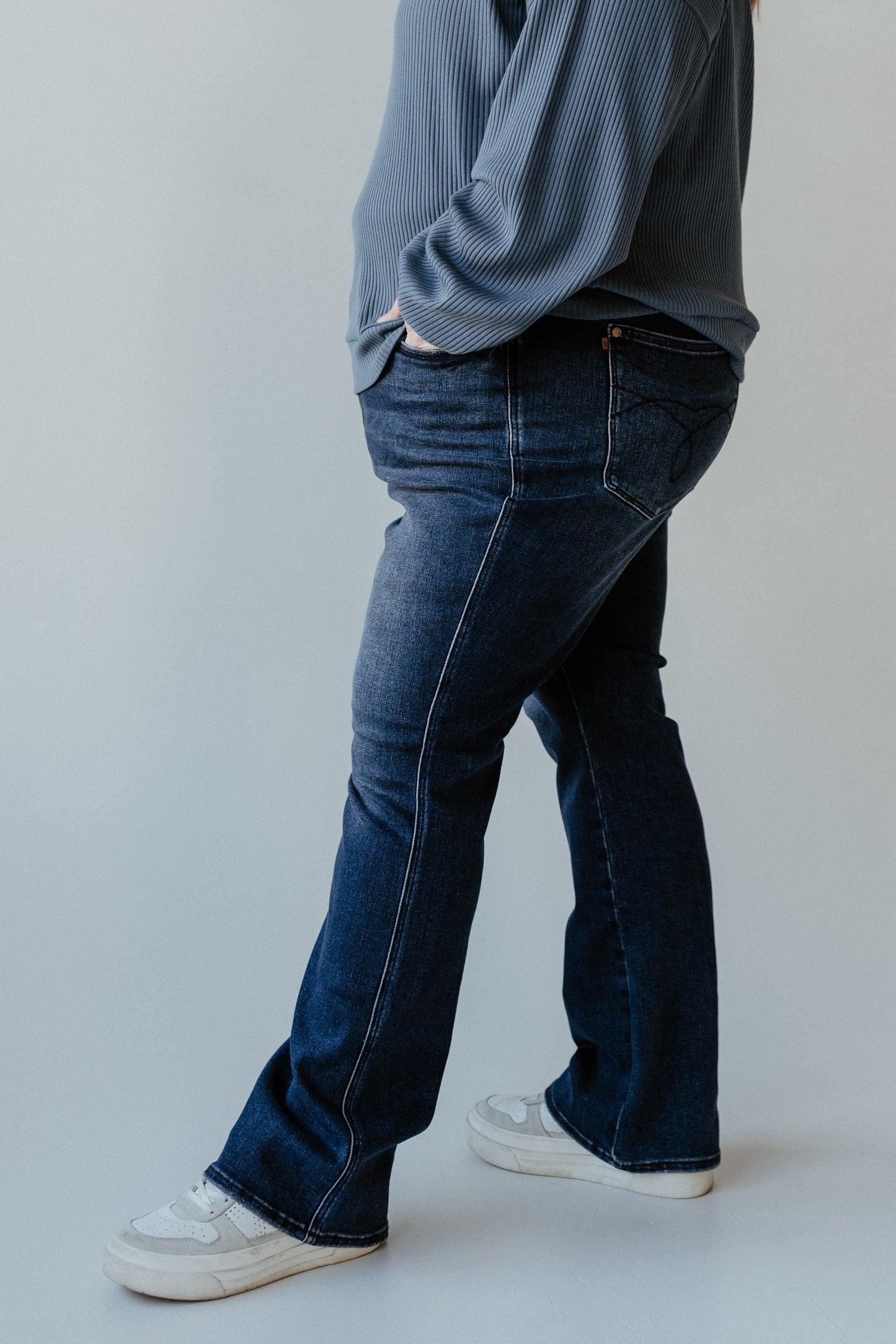 A person wearing a gray ribbed sweater, dark blue bootcut jeans with an embroidered pocket, and white sneakers stands sideways with hands in pockets against a plain background.