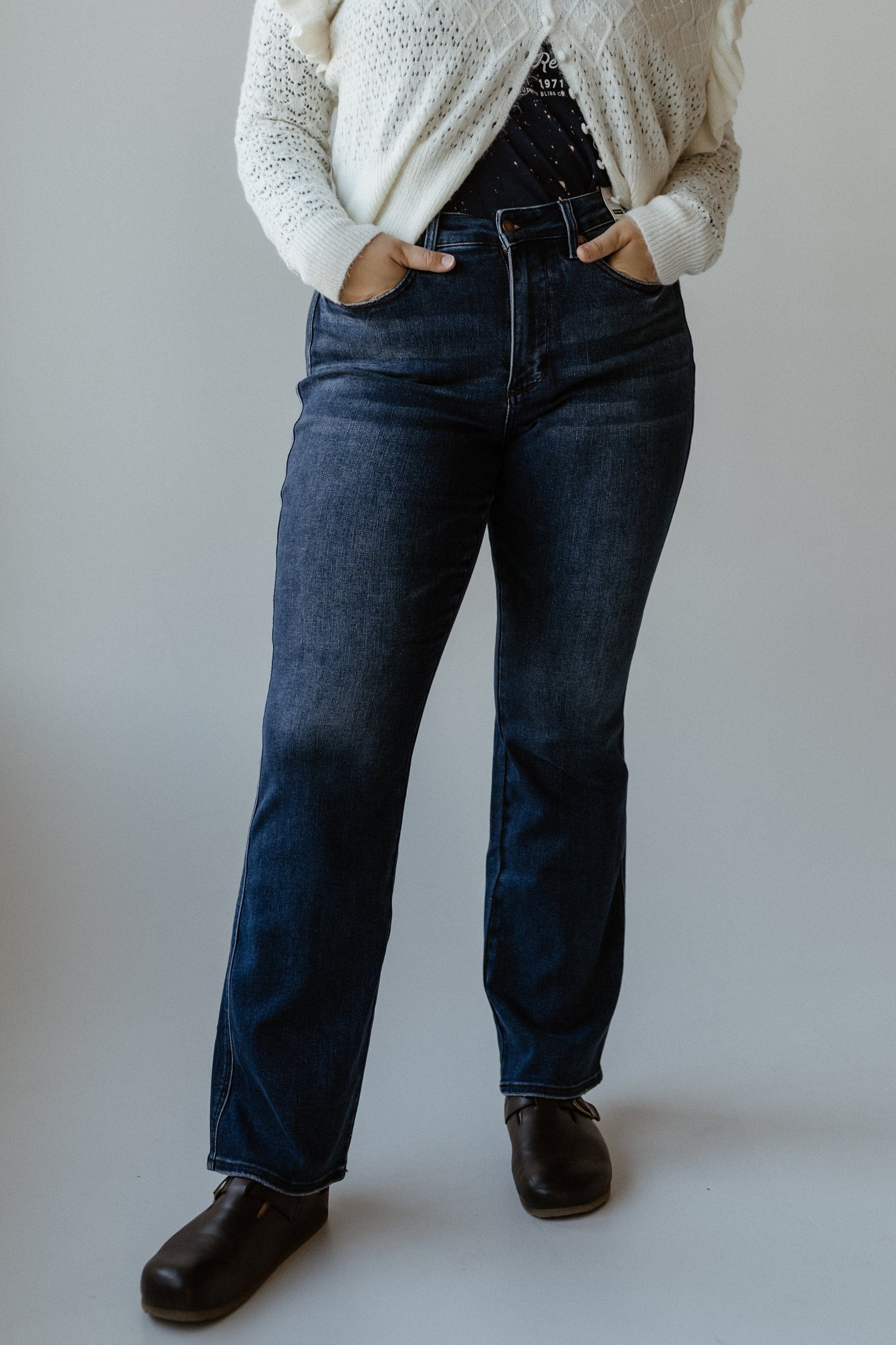 A person wearing blue bootcut jeans, a white sweater, and black shoes stands with hands in embroidered pockets against a plain background.