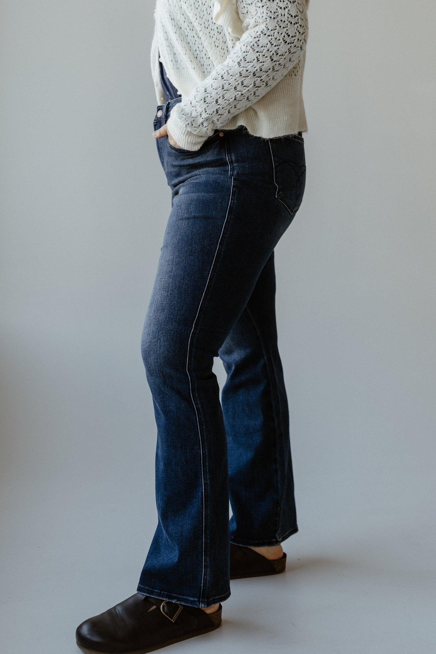 A person wearing a white lace sweater, bootcut jeans with an embroidered pocket, and black shoes stands in a neutral pose against a plain background.