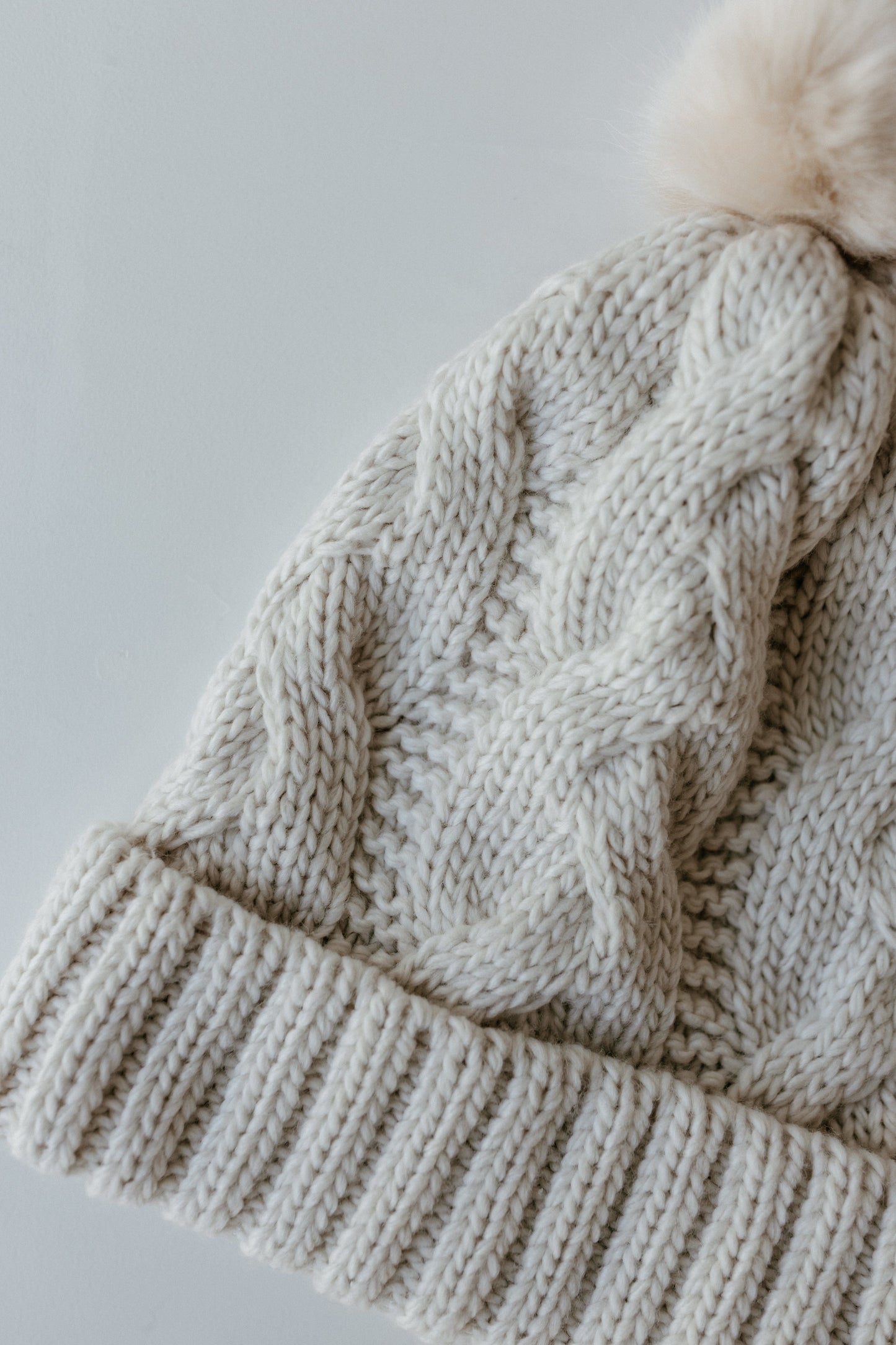 A cream-colored knitted beanie with a ribbed cuff and pom-pom detail, this cozy winter accessory perfectly complements your seasonal wardrobe.